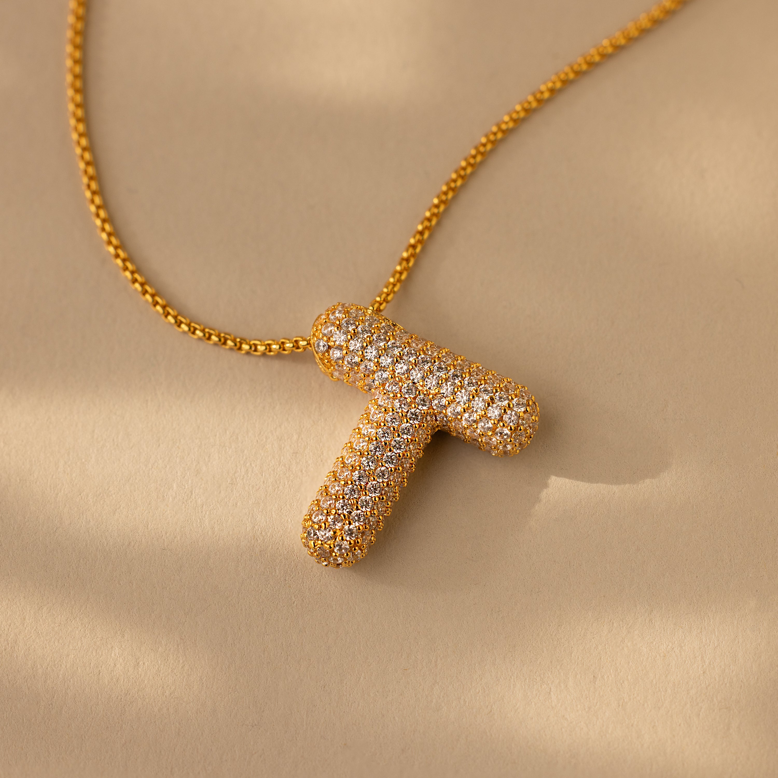 The Pave Puff Letter Necklace in 18K Gold features a gold "T" letter charm with pave diamonds, shown on a beige background.