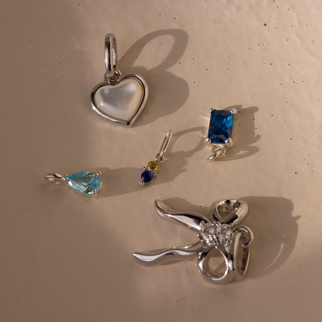 Several different silver charms including the dainty duo birthstone charm, teardrop birthstone charm, emerald birthstone charm, bow charm and pearl heart charm
