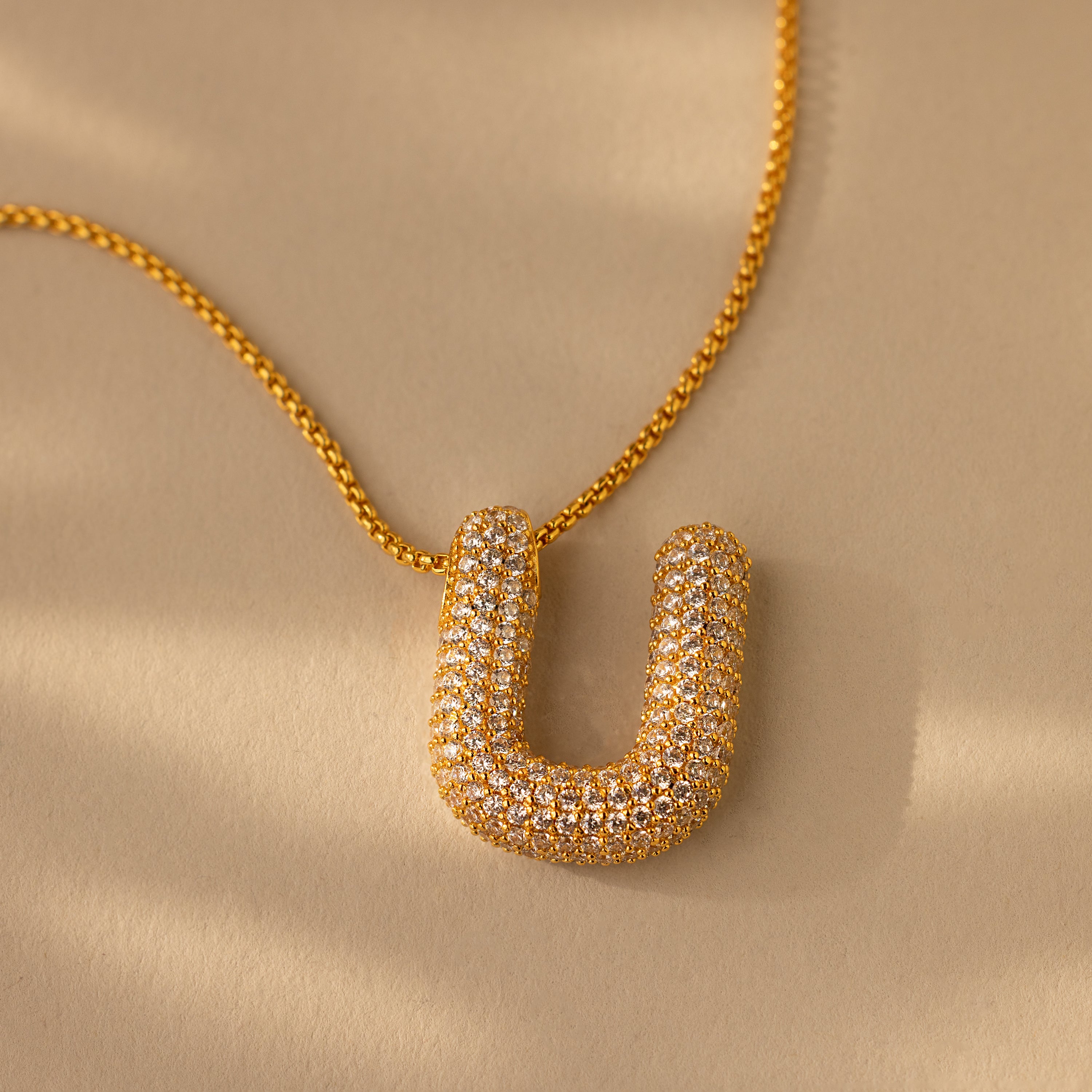 The Pave Puff Letter Necklace in 18K Gold features a U-shaped pendant encrusted with pavé diamonds, elegantly displayed against a beige background.