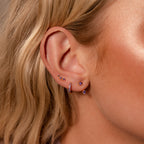 Close-up of a woman's ear wearing the Opal Earring Trio Set—multiple purple birthstone and huggie hoop earrings—framed by wavy blonde hair.