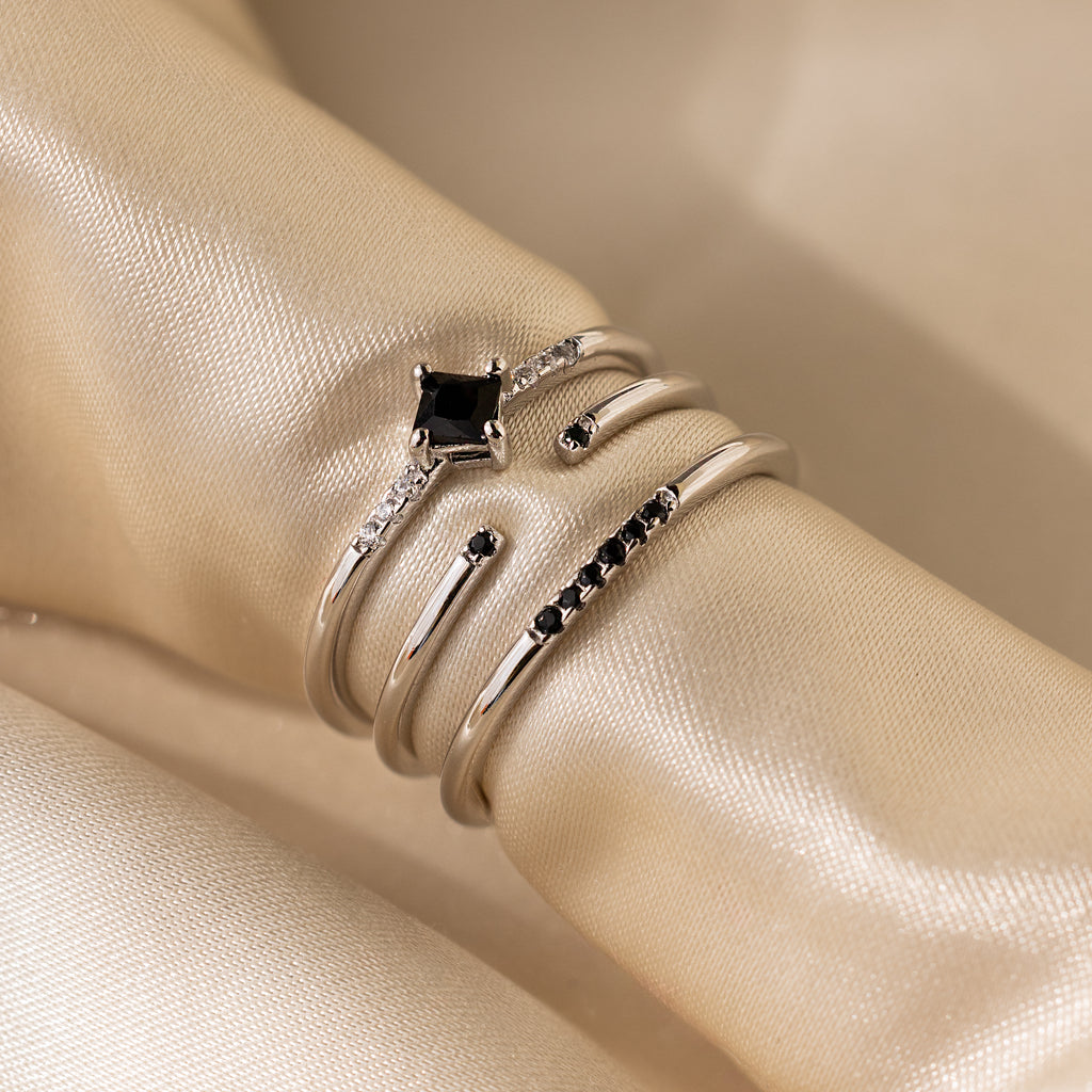 The Black Abyss Ring Set includes three silver bands with black and clear stones, showcased on silky beige fabric—perfect for fans of dainty rings.