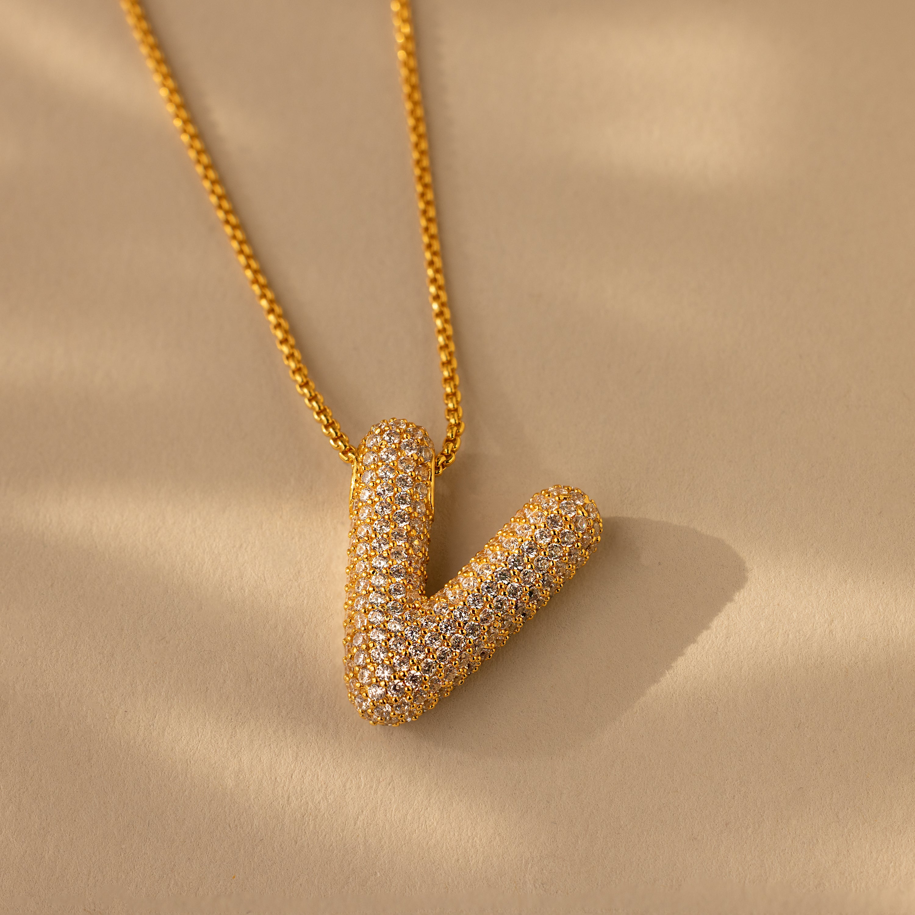 The Pave Puff Letter Necklace in 18K Gold features a V-shaped pendant with pave diamonds, echoing elegant design, displayed on a beige background.