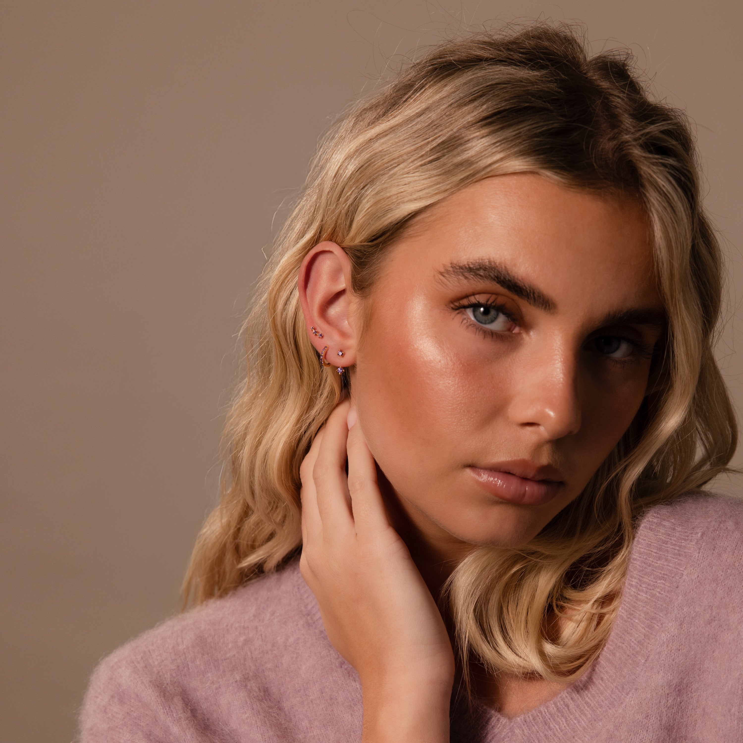 Woman with wavy blonde hair in a lilac sweater touches her ear, which is adorned with a pavé huggie hoop from the Alexandrite Earring Trio Set in gold, set against a neutral background.