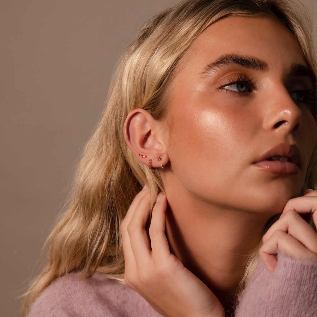Close-up of a woman with blonde hair wearing the Birthstone Earring Trio Set and a soft pink sweater, gently touching her ear.