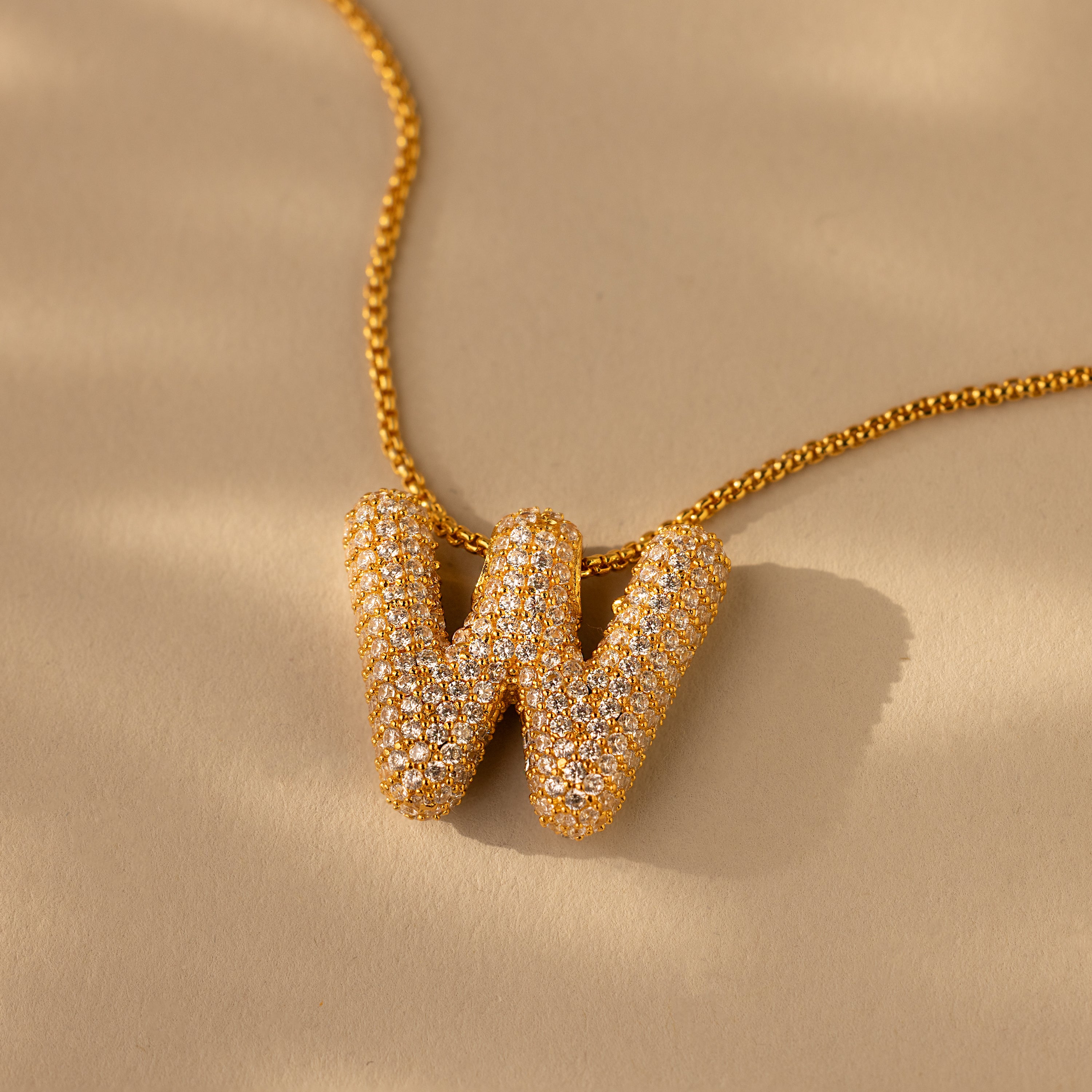 The Pave Puff Letter Necklace in 18K Gold features a sparkling gold W letter pendant with pave diamonds, shown on a beige background.