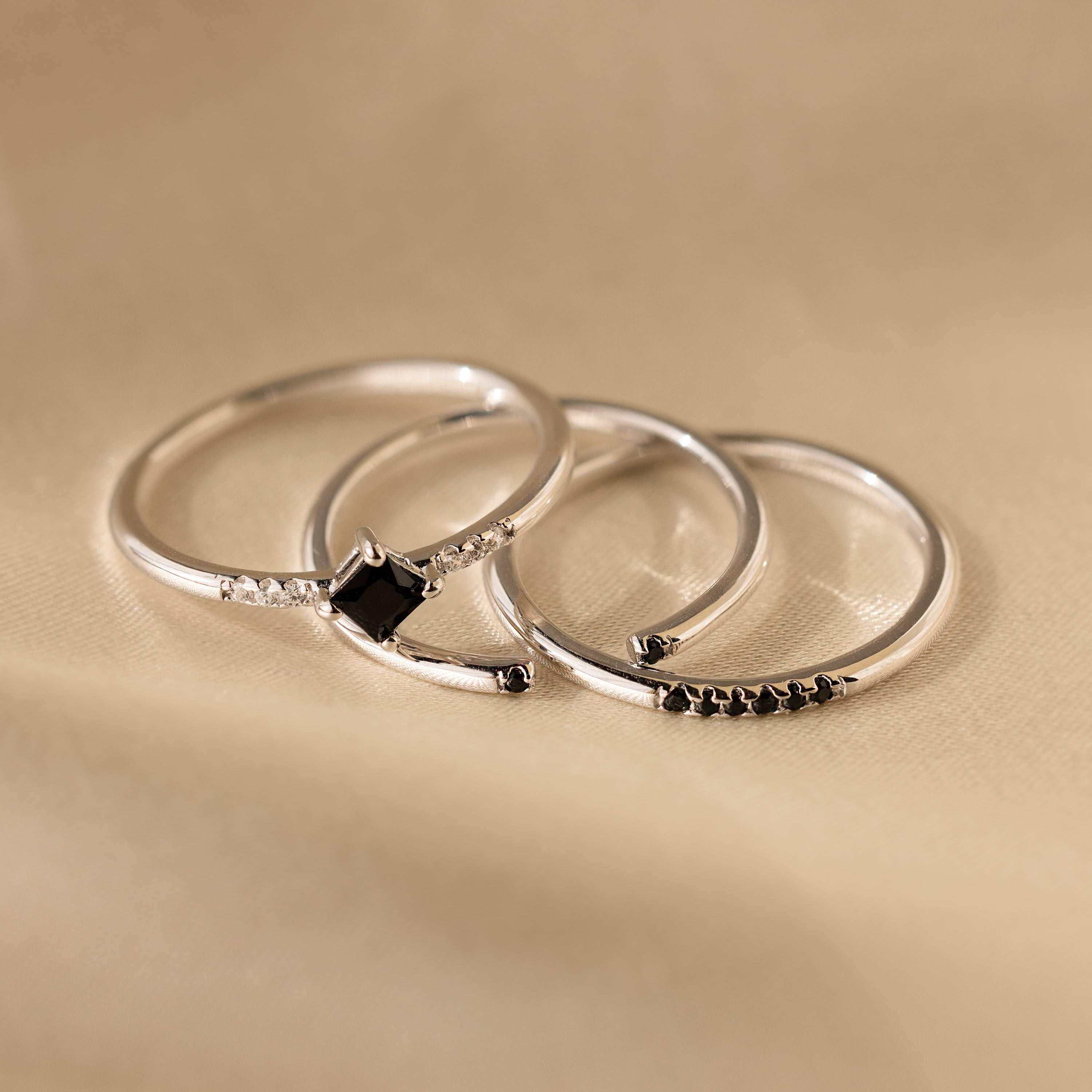 Three delicate silver bands from the Black Abyss Ring Set in Silver, each with a small black gemstone, displayed on a soft beige fabric background.