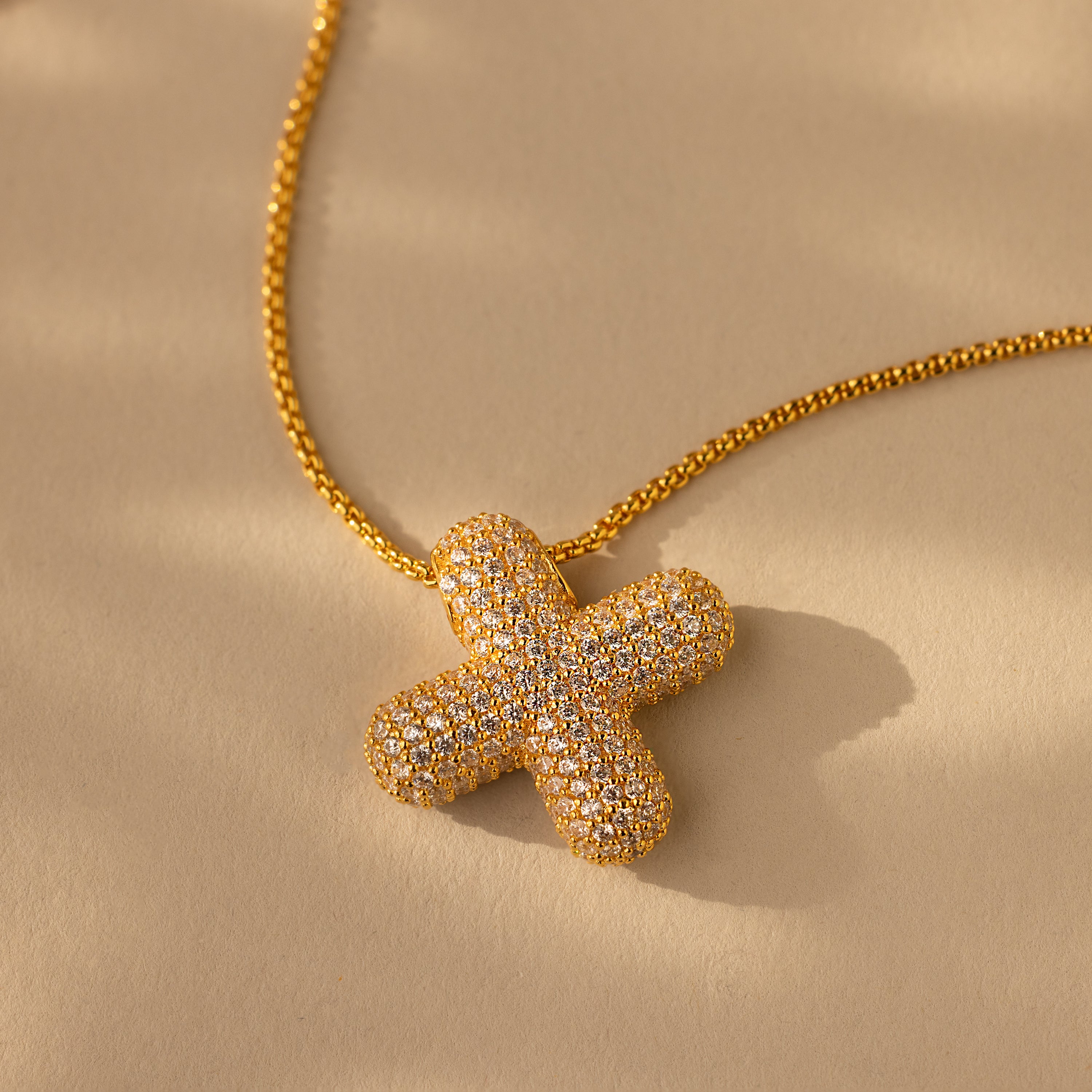 Showcased on a beige background, the Pave Puff Letter Necklace in 18K Gold features a textured, diamond-studded letter pendant with sparkling pave diamonds.