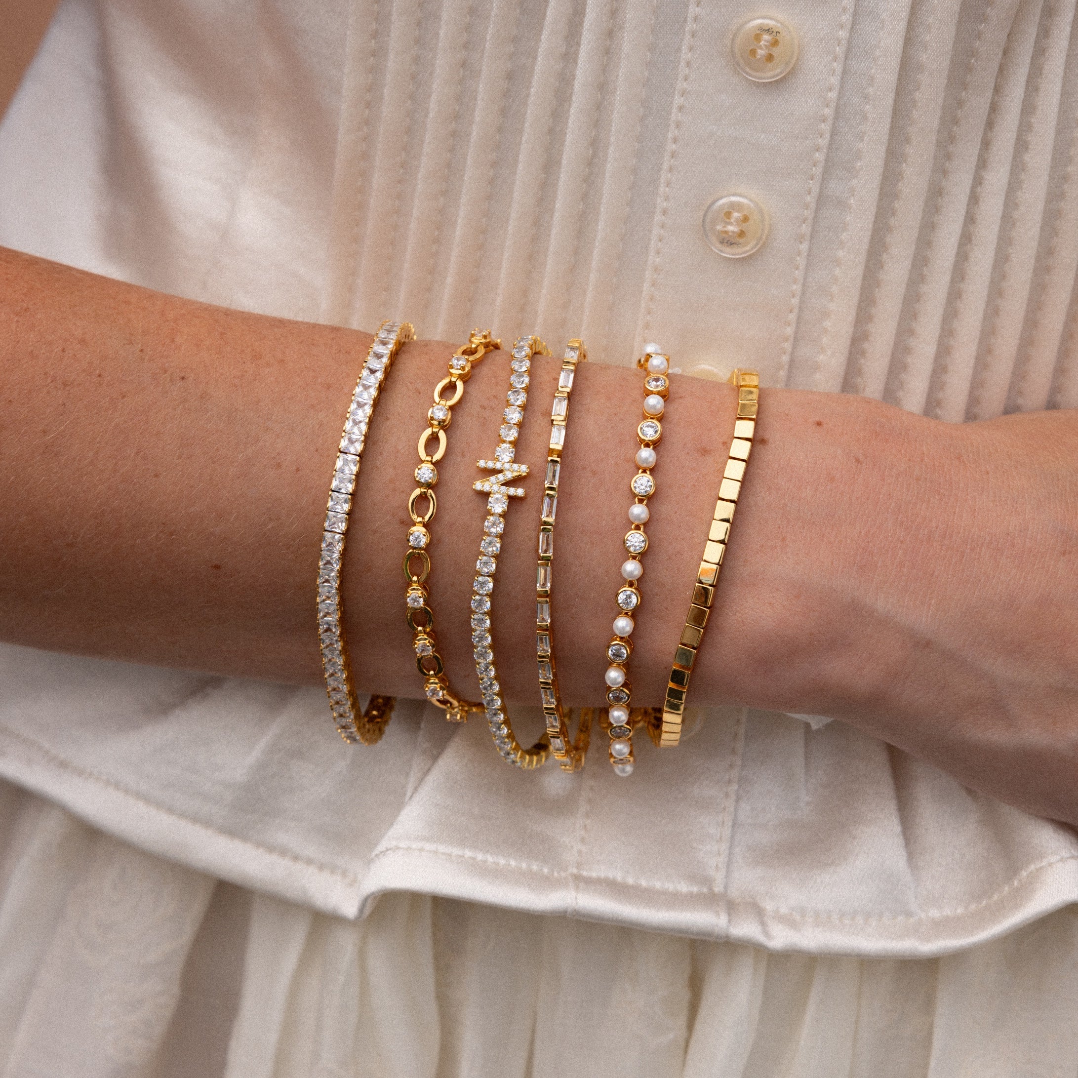 A person wearing a white blouse and five gold bracelets, including the Princess Diamond Tennis Bracelet with sparkling princess-cut diamonds.