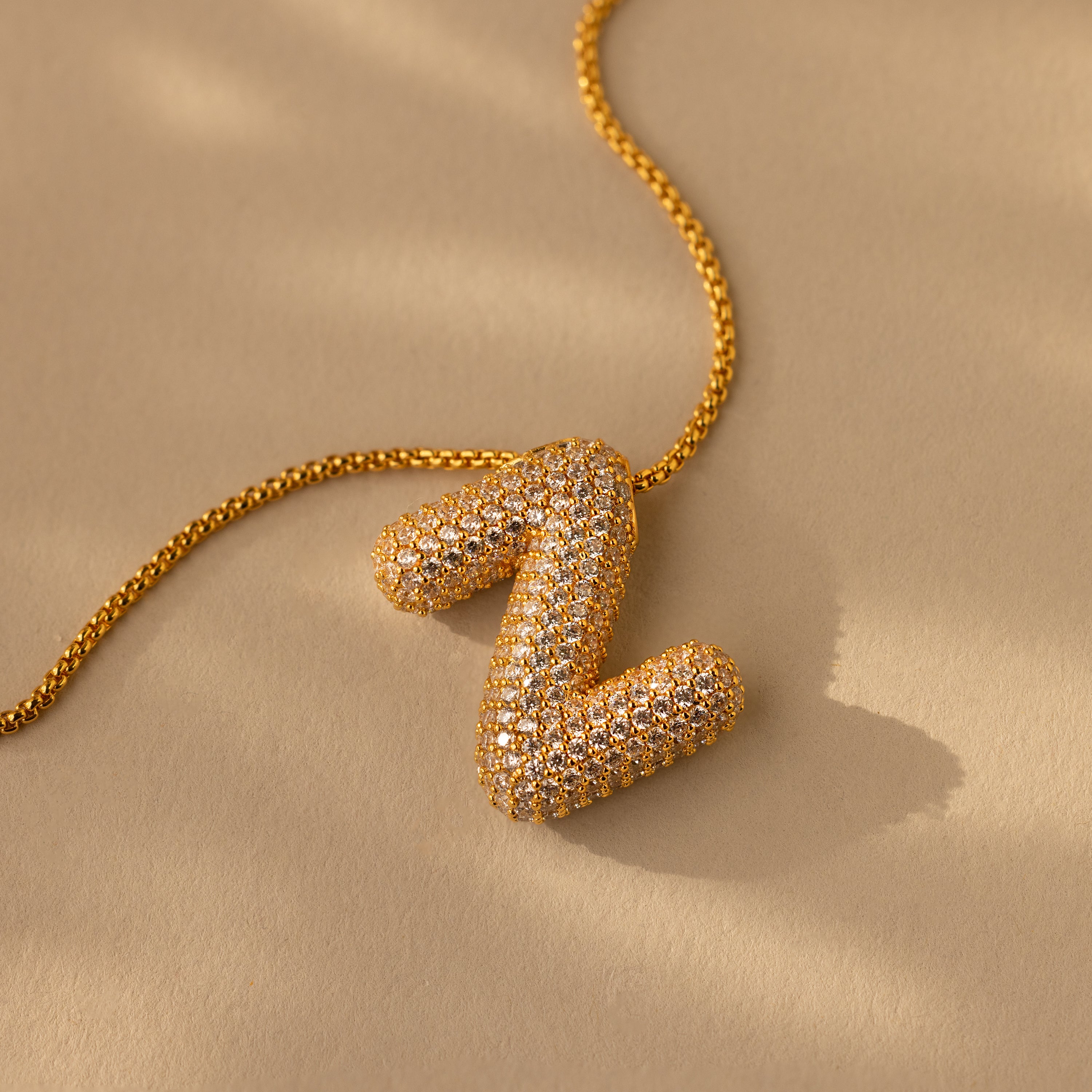 The Pave Puff Letter Necklace in 18K Gold features a diamond-encrusted gold letter Z charm, showcased on a beige surface with soft shadows.