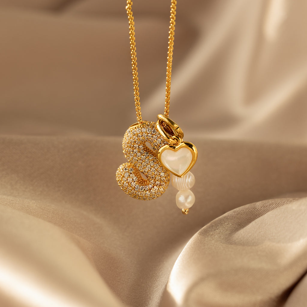 The Pave Puff Letter Necklace showcases a crystal-studded “S” pendant, a gold heart charm, and two pearl-like beads, elegantly arranged on silky beige fabric.
