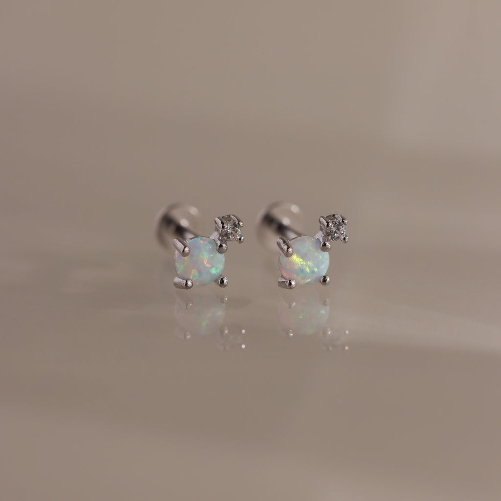 Studio shot of a pair of silver flatback stud earrings, each set with a round opal gemstone and a small diamond accent on a reflective surface.