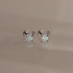 Studio shot of a pair of silver flatback stud earrings, each set with a round opal gemstone and a small diamond accent on a reflective surface.