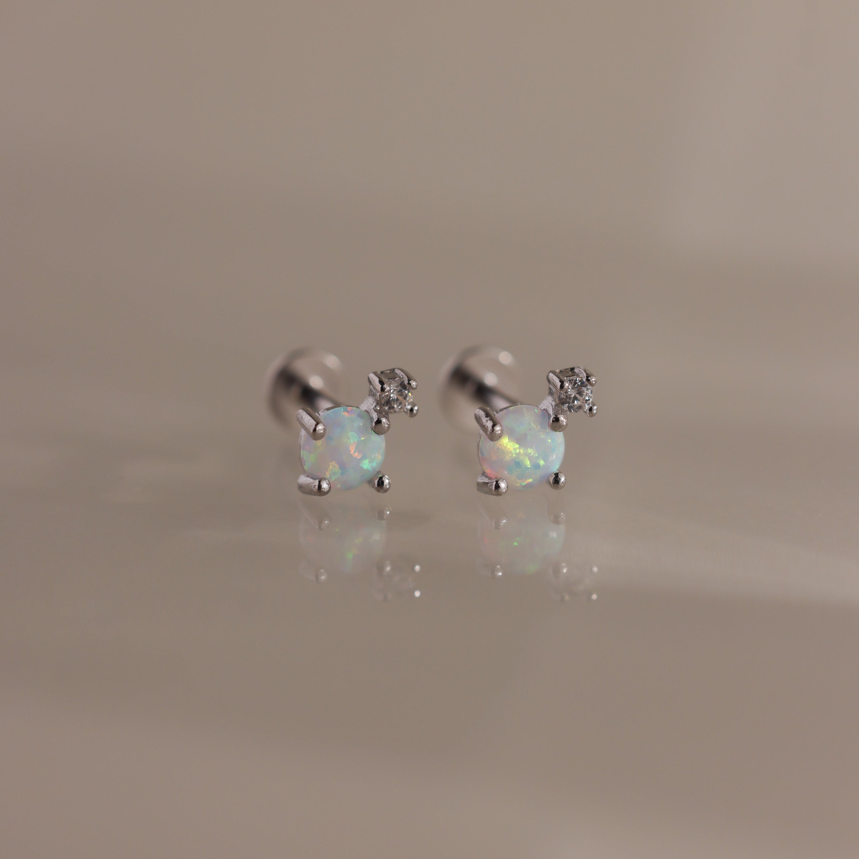 Studio shot of a pair of silver flatback stud earrings, each set with a round opal gemstone and a small diamond accent on a reflective surface.
