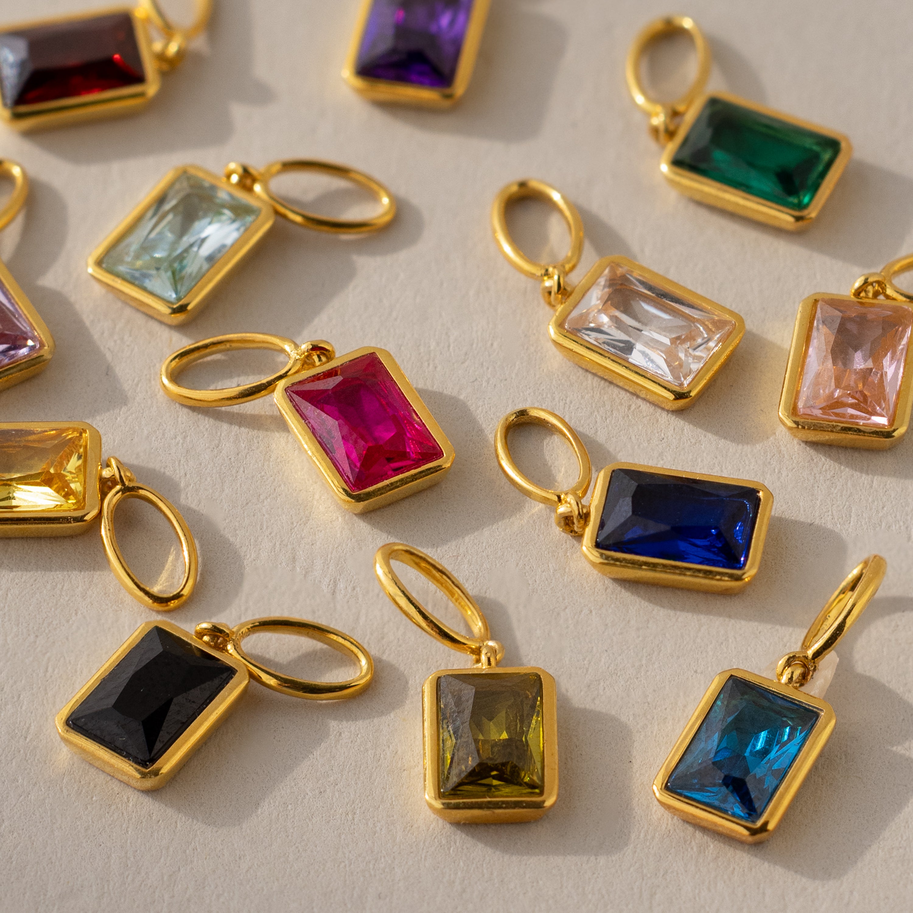 Multiple emerald-cut birthstone charms in different colors arranged side-by-side. Ruby and Sapphire charms are at the center