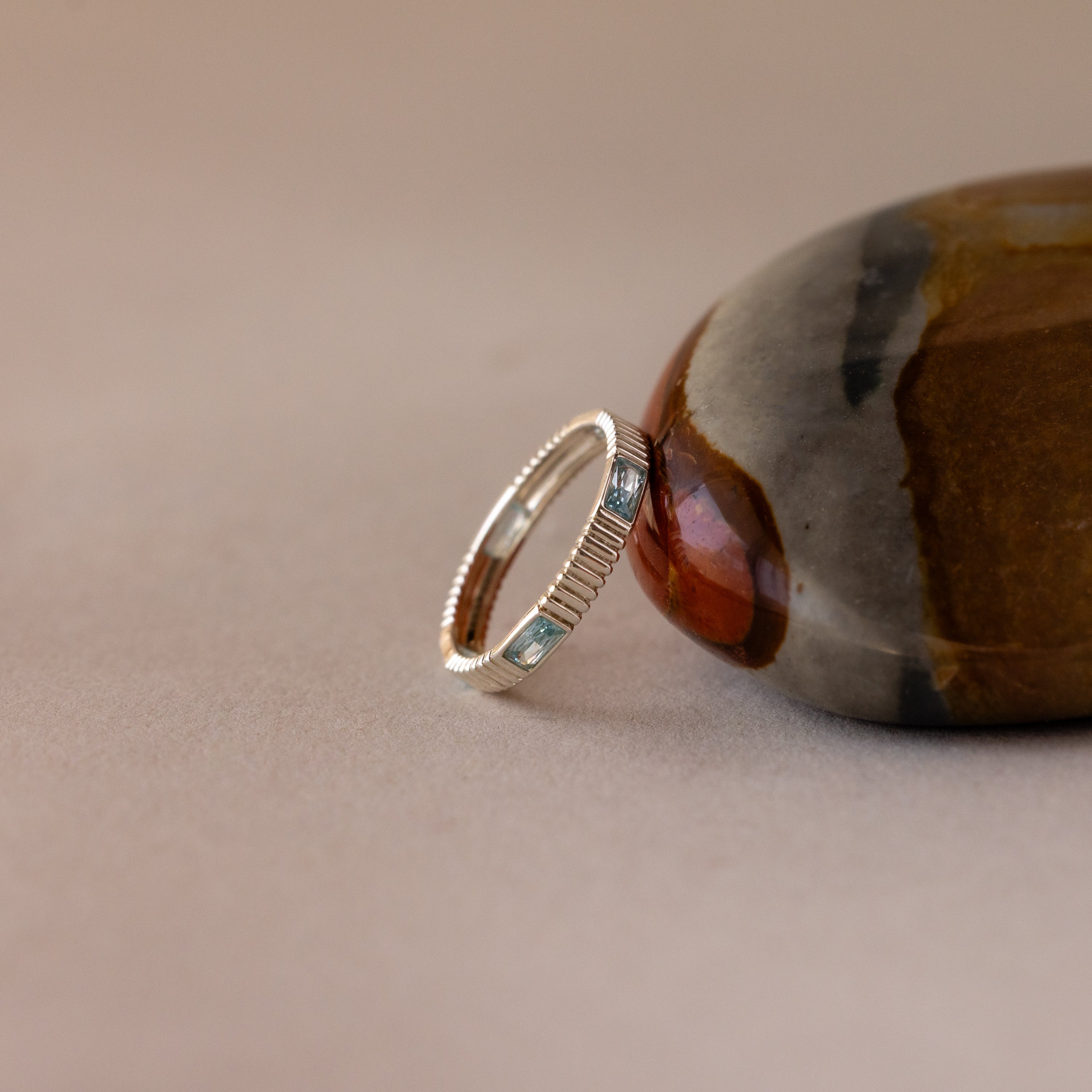 The Ribbed Baguette Birthstone Eternity Ring with blue rectangular stones is shown leaning against a smooth, multicolored stone on a beige surface.