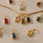 flat lay of three emerald-cut birthstone charms on a box chain, with more charms laying around them. In the center are Pink Tourmaline, Peridot and Sapphire stone charms