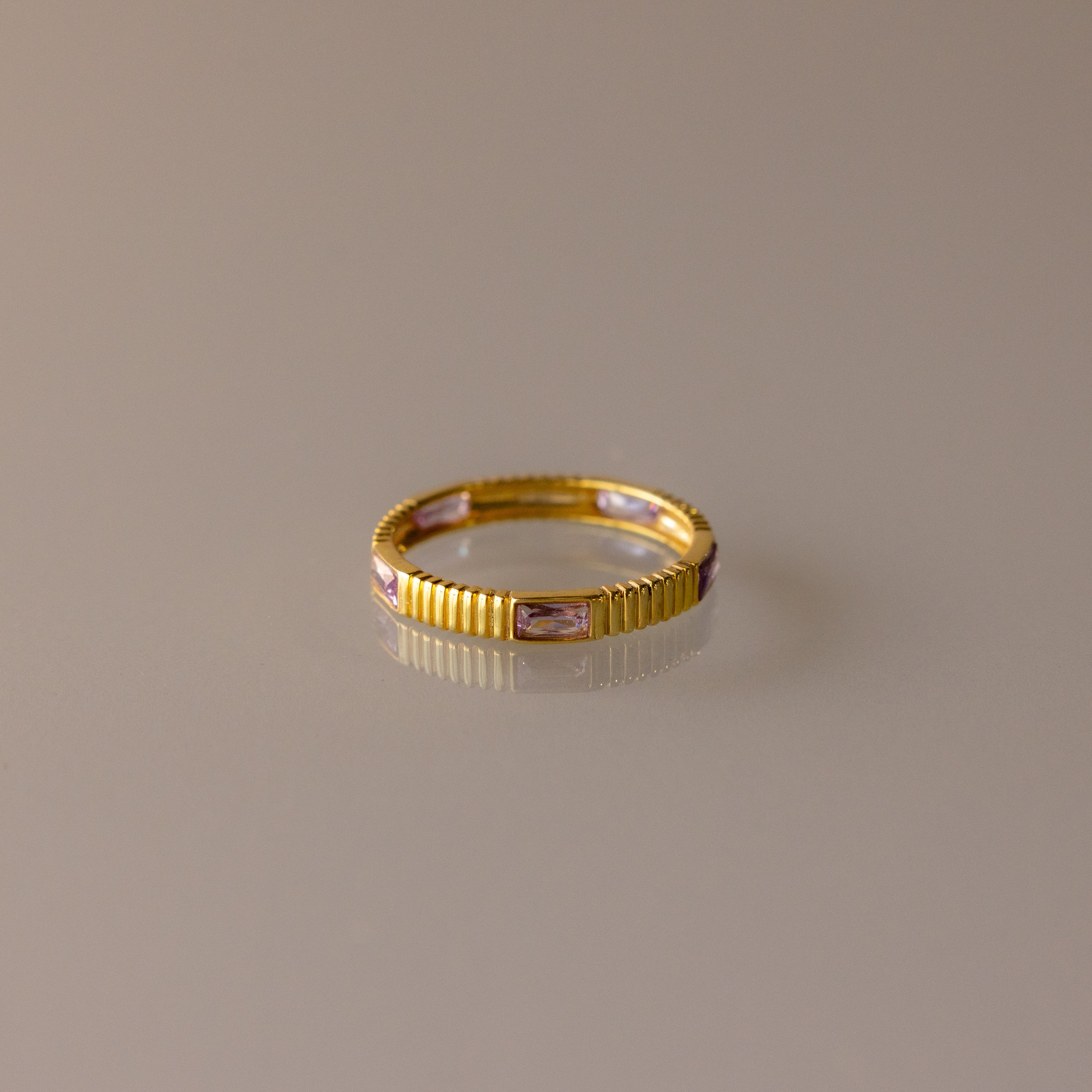 The Ribbed Baguette Alexandrite Eternity Ring features a slim gold band with textured ribbing and rectangular pink alexandrite gemstones set evenly around, shown against a neutral background.