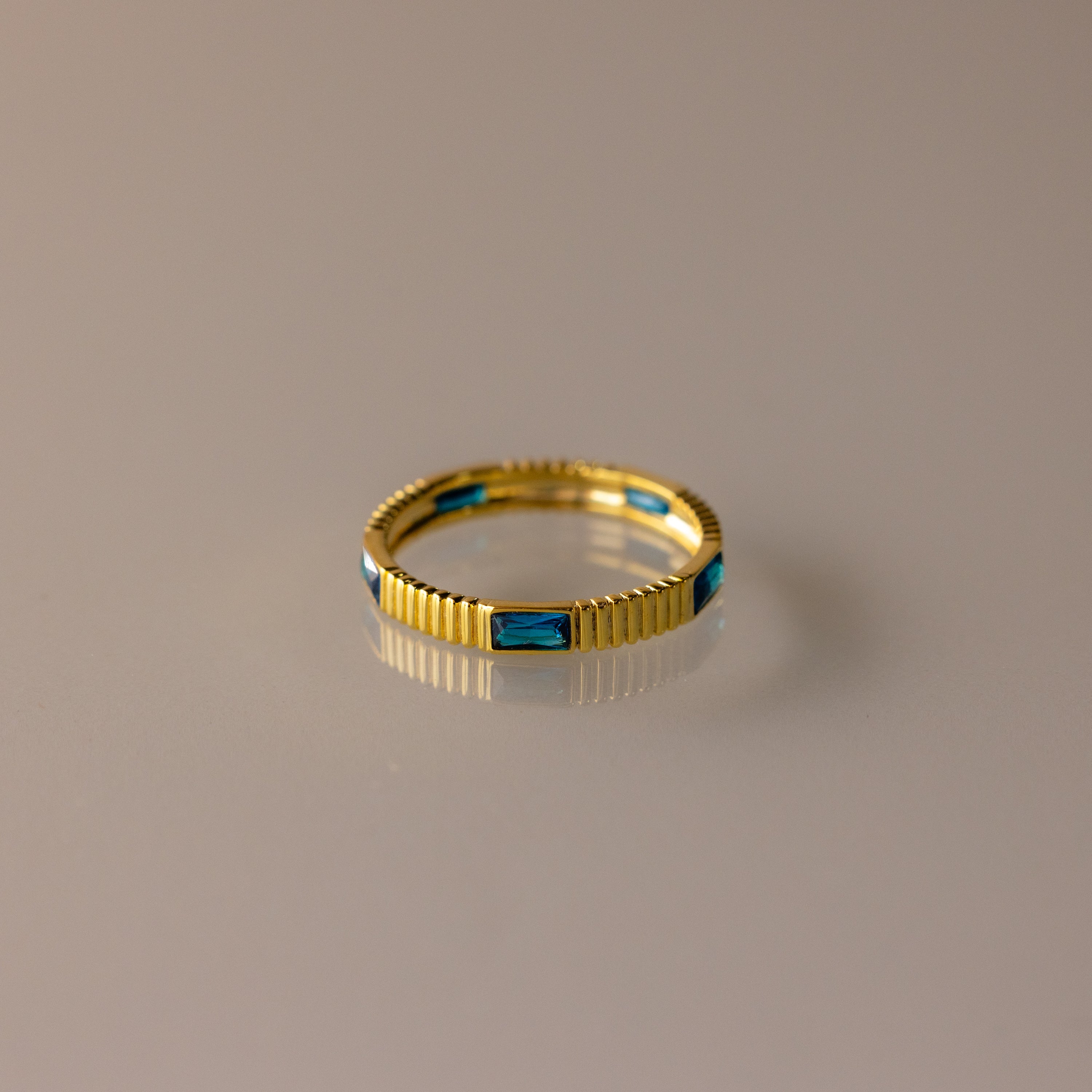 The Ribbed Baguette Blue Zircon Eternity Ring is a thin gold stackable ring with rectangular blue zircon stones, shown on a smooth, neutral surface.