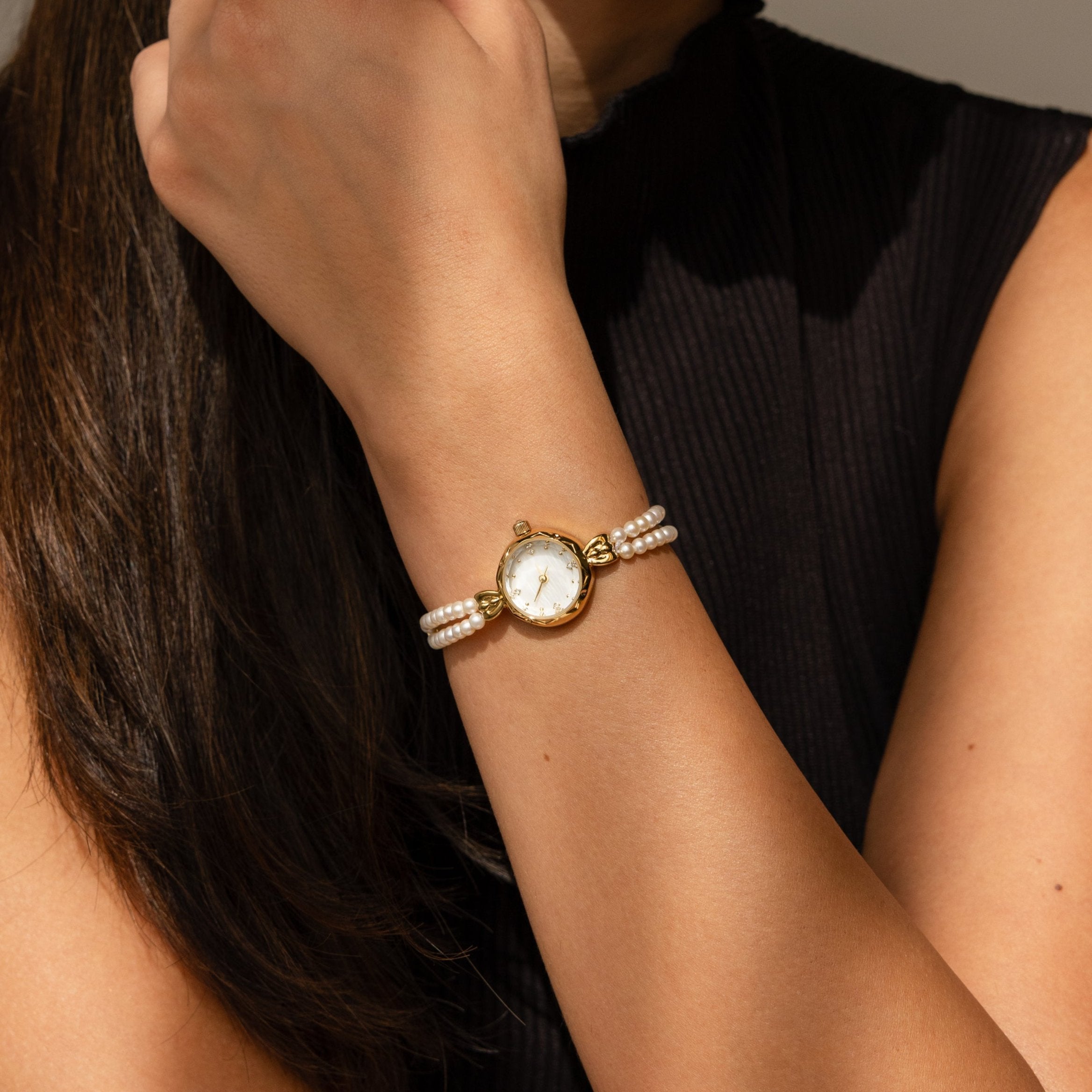 A woman in a sleeveless black top raises her hand to her face, showcasing the Beaded Pearl Timepiece on her wrist.
