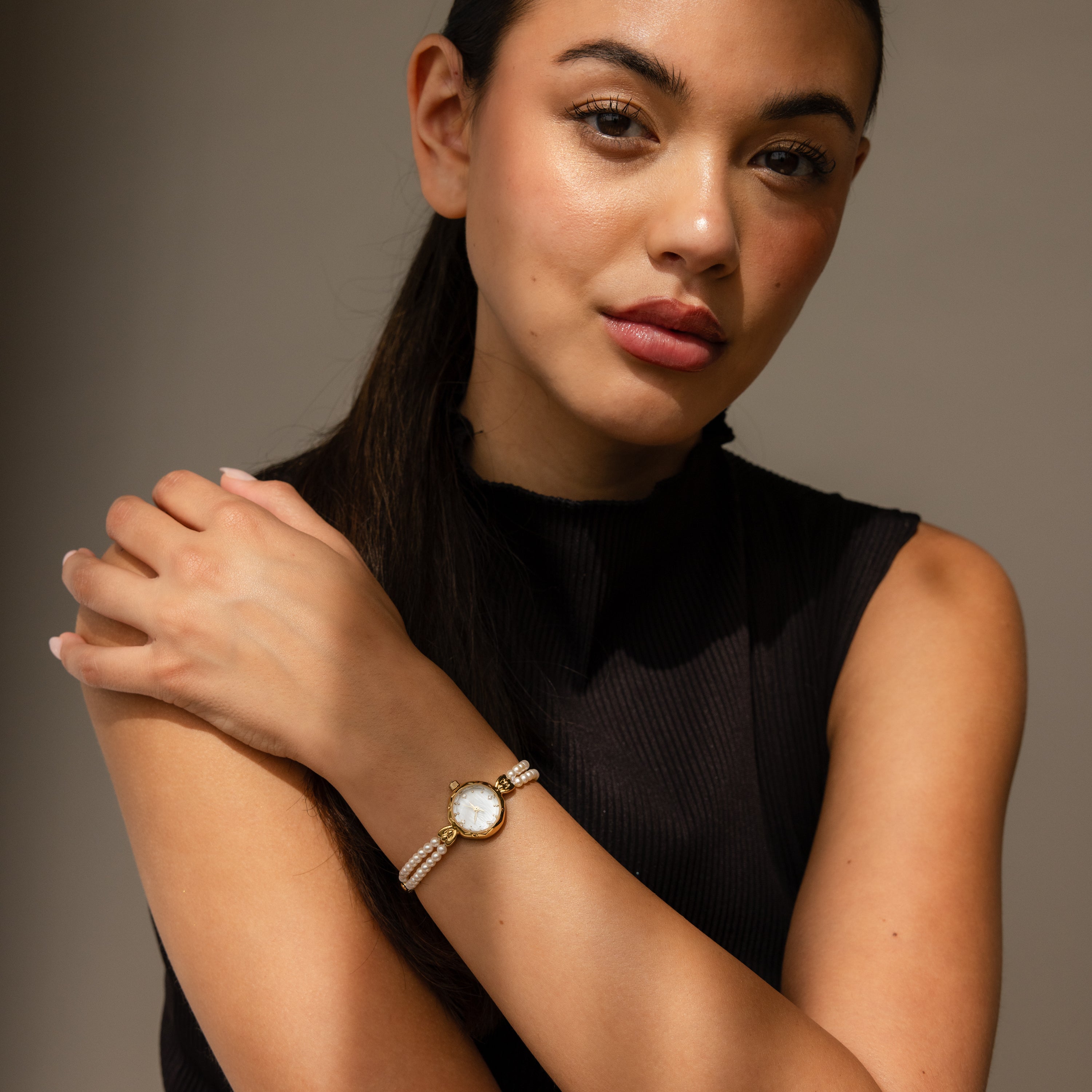 A woman in a black sleeveless top confidently models the Beaded Pearl Timepiece in 18K Gold, glancing at the camera.