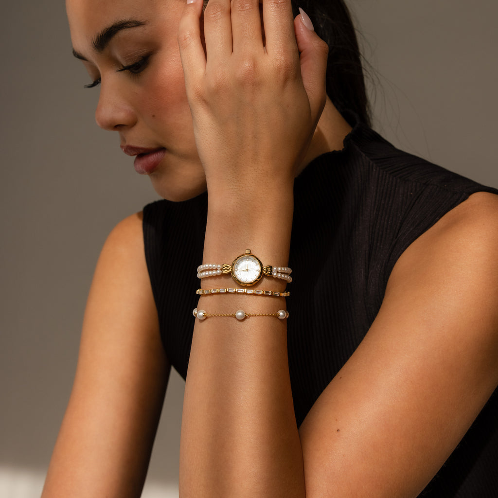 A woman thoughtfully looks down, wearing the Beaded Pearl Timepiece—a chic black sleeveless top pairs elegantly with this classic watch and pearl bracelet combo.