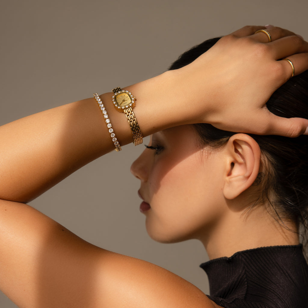 A woman in a black top wears the elegant Pave Blossom Timepiece, featuring a gold bracelet on her wrist, as she rests her hand on her head against a neutral background.
