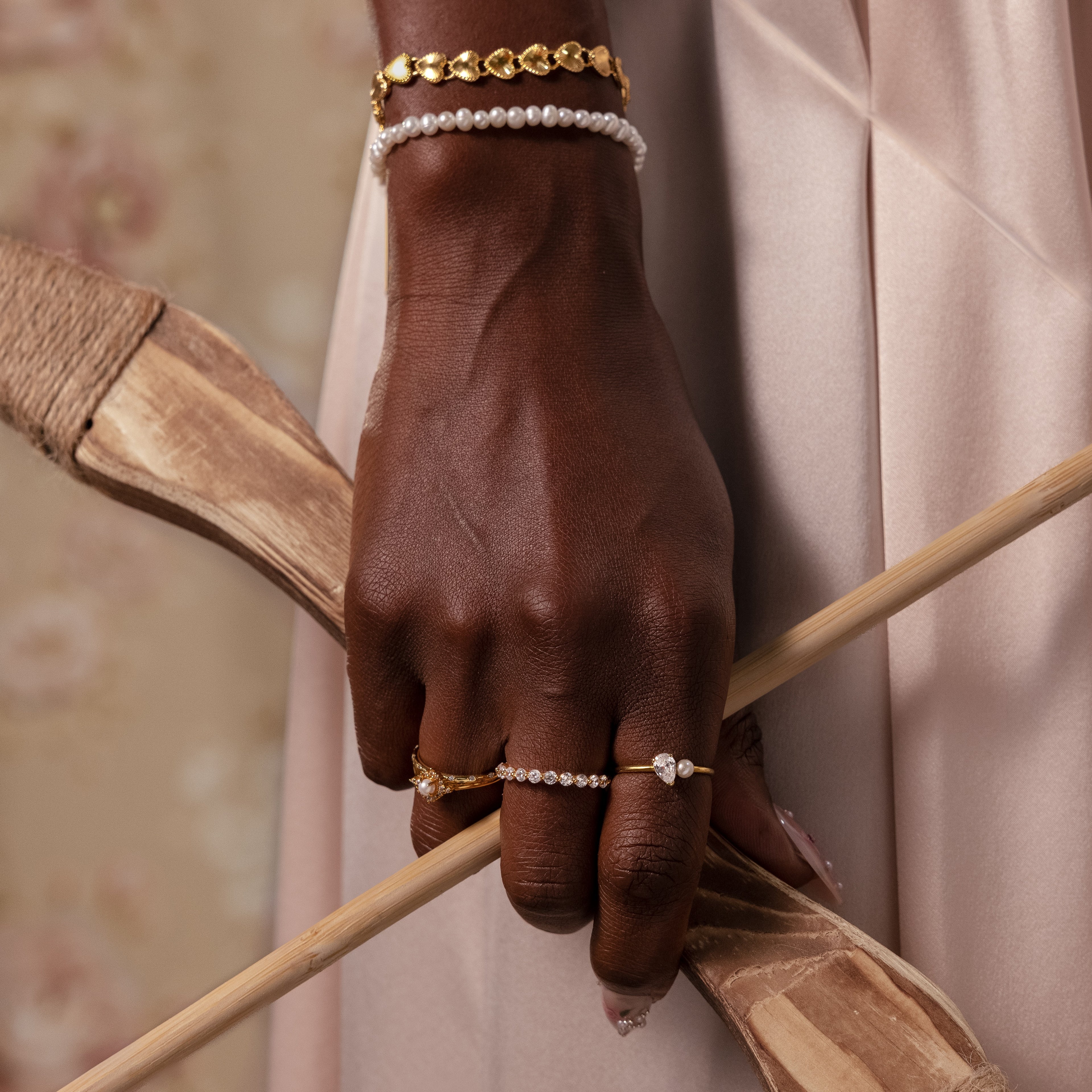 A hand adorned with elegant jewelry, including the Venus Pearl Bracelet, holds wooden sticks against a soft beige background.
