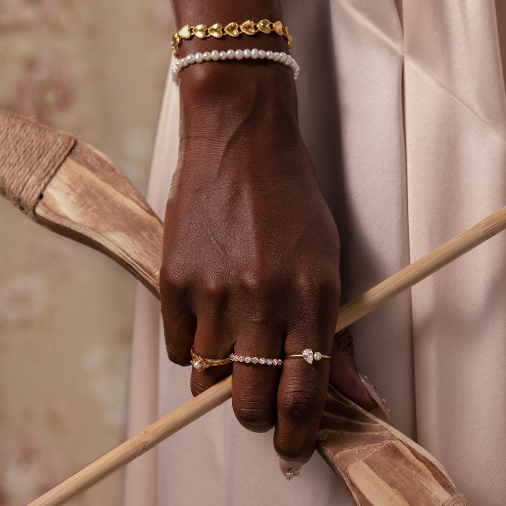 A hand wearing the Ribbed Heart Link Bracelet holds wooden objects against a soft beige background, showcasing the bracelet’s vintage elegance and romantic style.