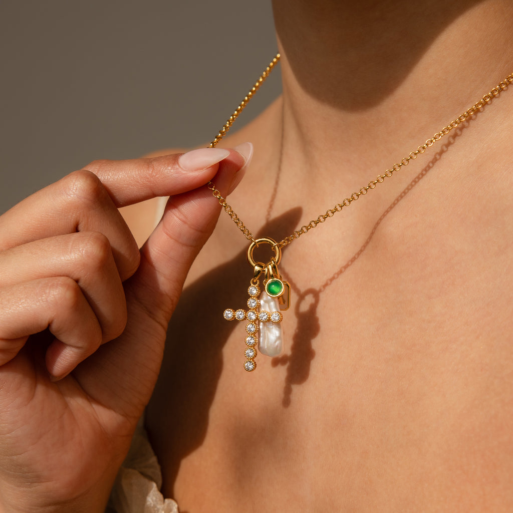 A person holds a gold necklace with gemstone charms—a jeweled cross, a pearl, and a green Signet Birthstone Charm—showcasing elegant personalized jewelry near their neck.