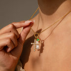 A person holds a gold necklace with gemstone charms—a jeweled cross, a pearl, and a green Signet Birthstone Charm—showcasing elegant personalized jewelry near their neck.