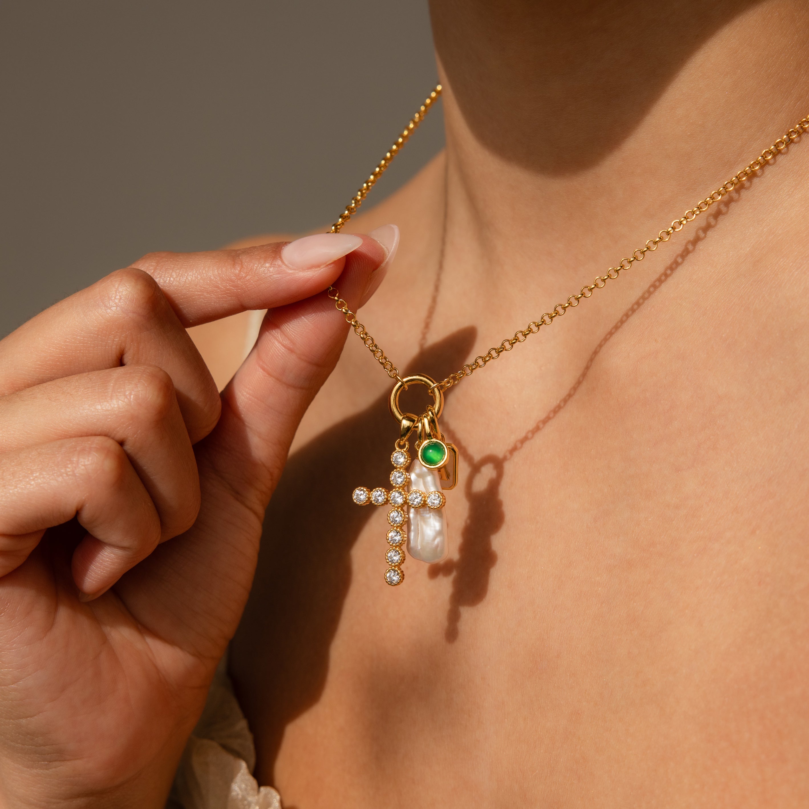 A person holds a gold necklace with gemstone charms—a jeweled cross, a pearl, and a green Signet Birthstone Charm—showcasing elegant personalized jewelry near their neck.