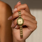 A hand with manicured nails holds the Pave Blossom Timepiece, an elegant womens watch featuring a gold case with a bezel adorned in green CZ gemstones.