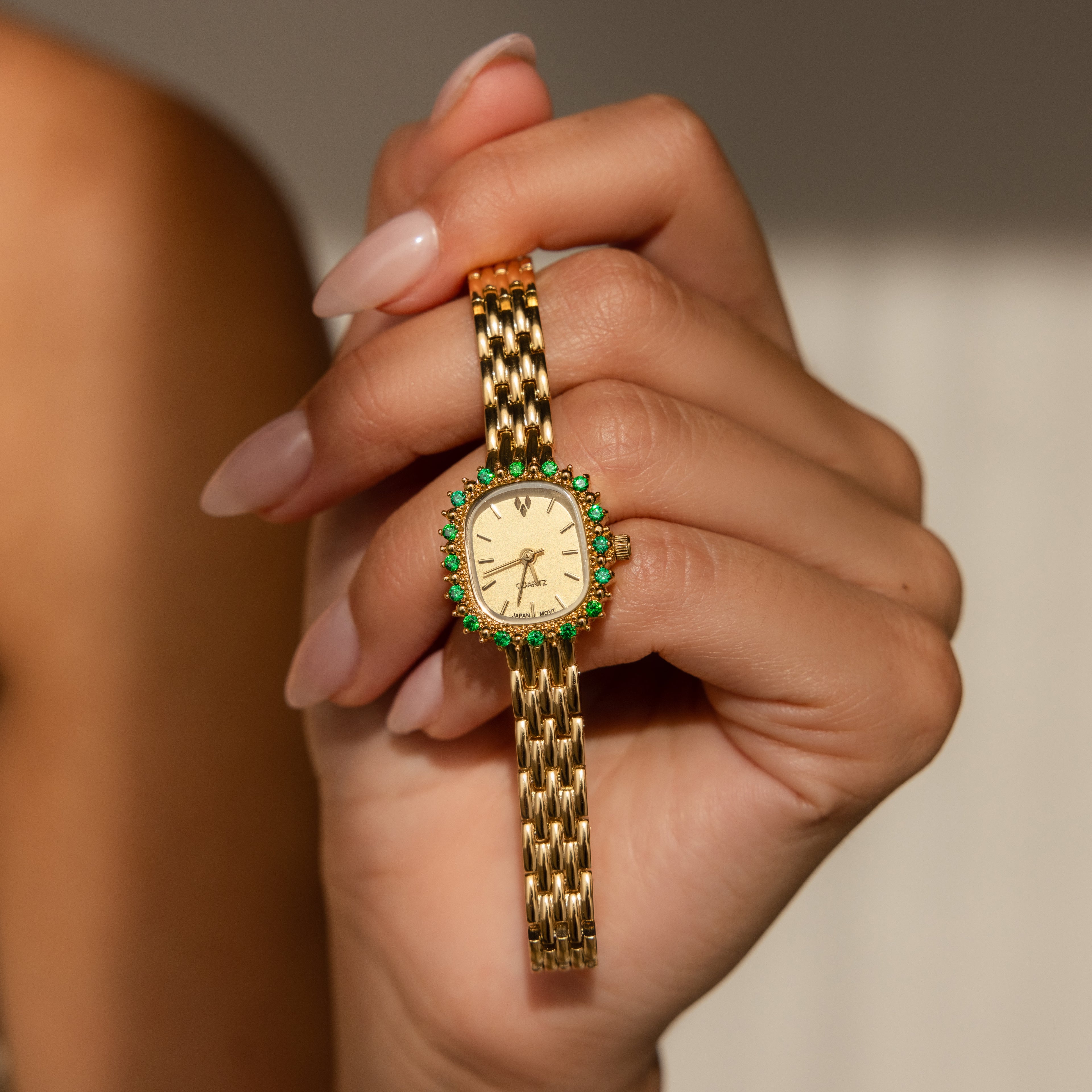 A hand with manicured nails holds the Pave Blossom Timepiece, an elegant womens watch featuring a gold case with a bezel adorned in green CZ gemstones.