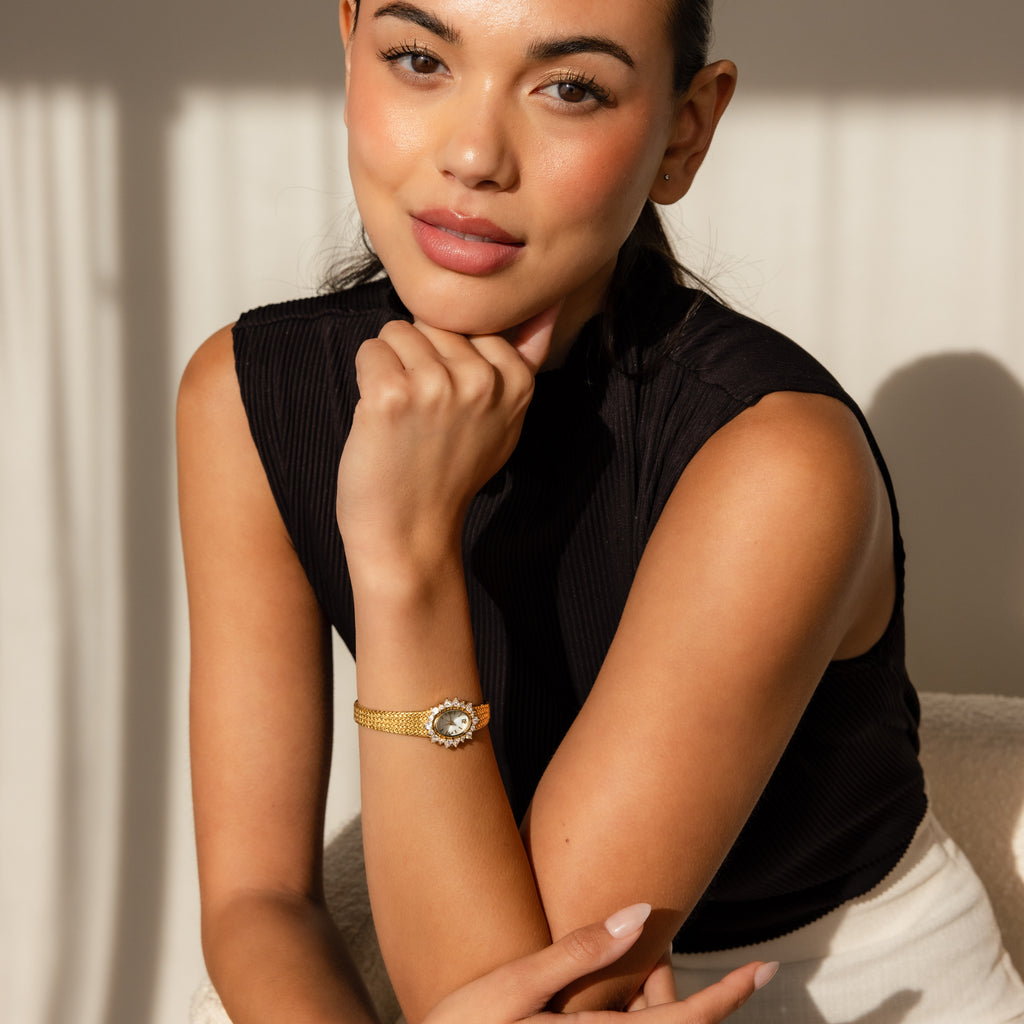 A woman in a black sleeveless top poses with her chin on her hand, showcasing the elegant Pave Heart Timepiece—a watch adorned with heart-shaped gemstones.