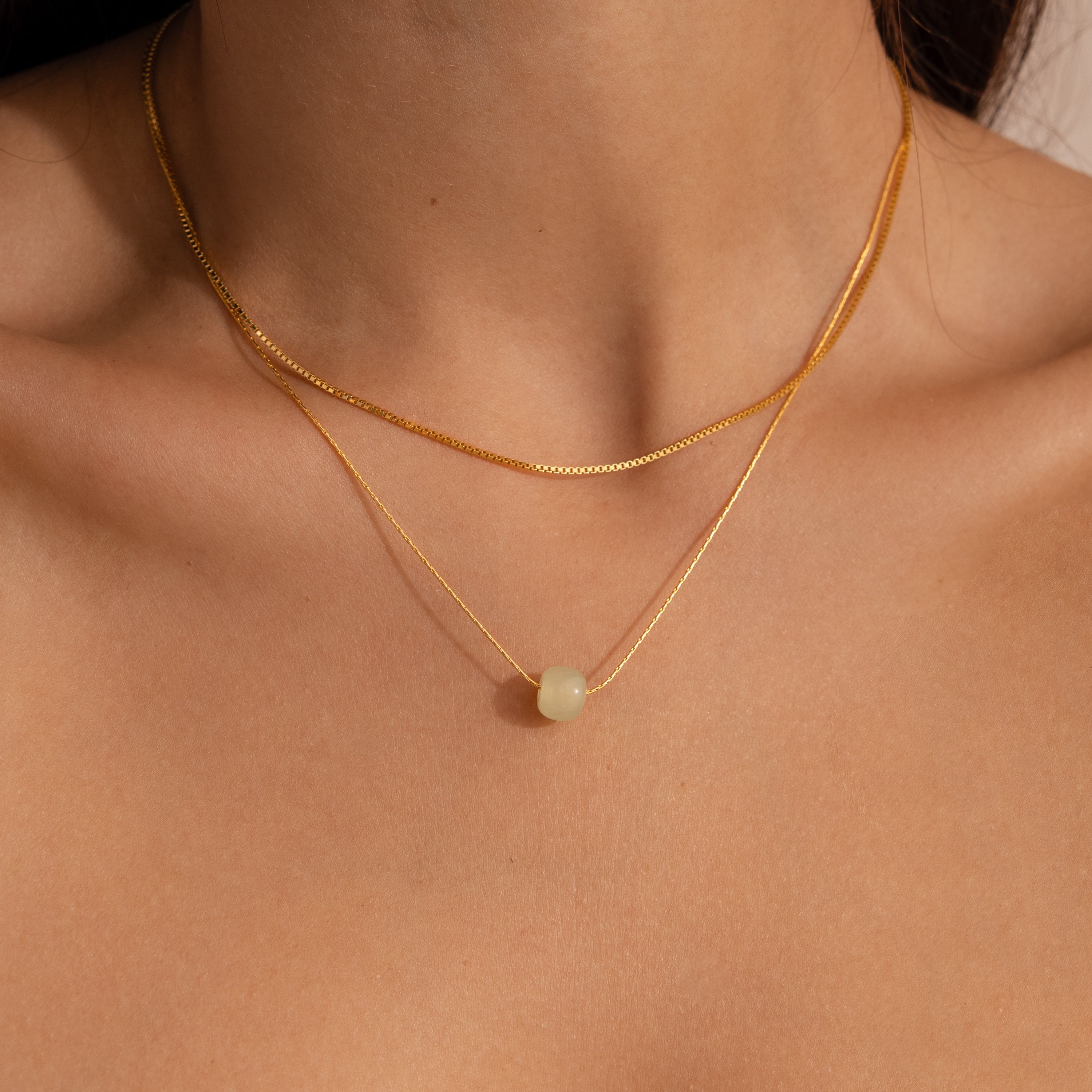 Close-up of model wearing a delicate gold chain necklace featuring a single round green jade bead, layered with a dainty box chain necklace.