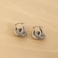 Duo Pave Link Hoops in White Gold: Elegant white gold hoop earrings feature intertwined, textured loops with a touch of sparkle, displayed on a soft beige background.