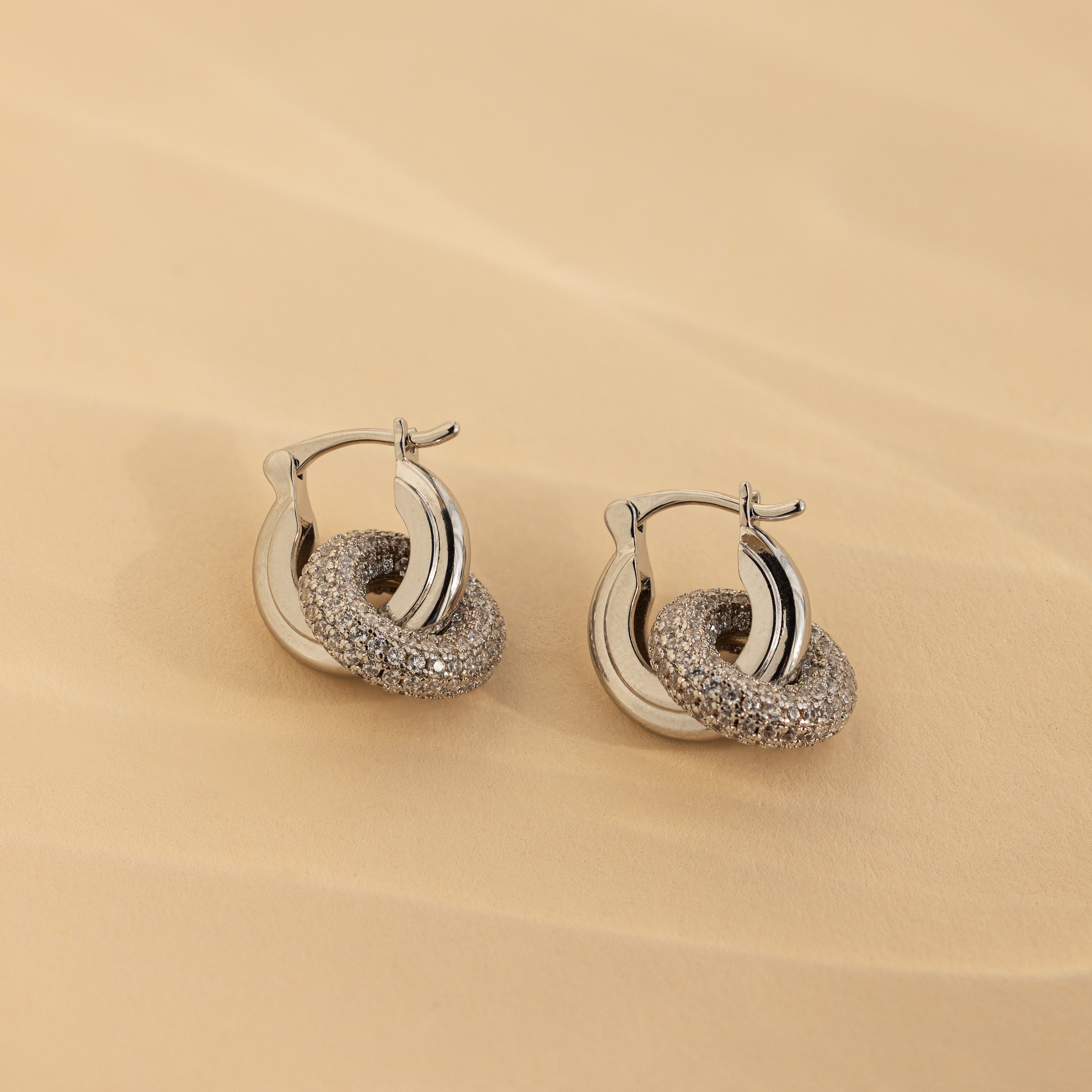 Duo Pave Link Hoops in White Gold: Elegant white gold hoop earrings feature intertwined, textured loops with a touch of sparkle, displayed on a soft beige background.