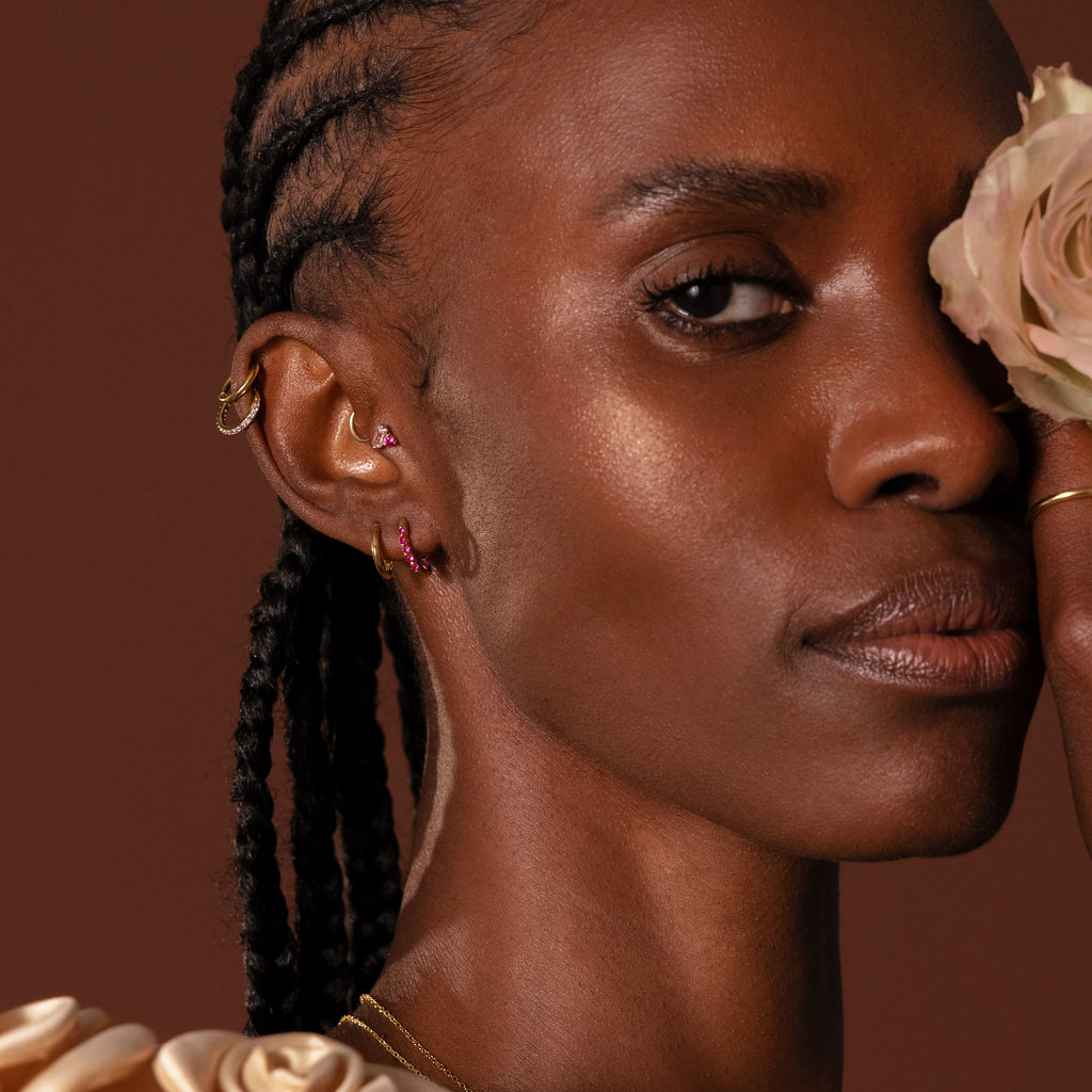 Woman with braided hair and multiple ear piercings wears Inside Out Ruby Huggies, holding a pale rose near her face against a brown background.