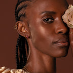 Woman with braided hair and gold jewelry, including Diamond Clicker Huggies, holds a cream rose to her face against a warm brown background.