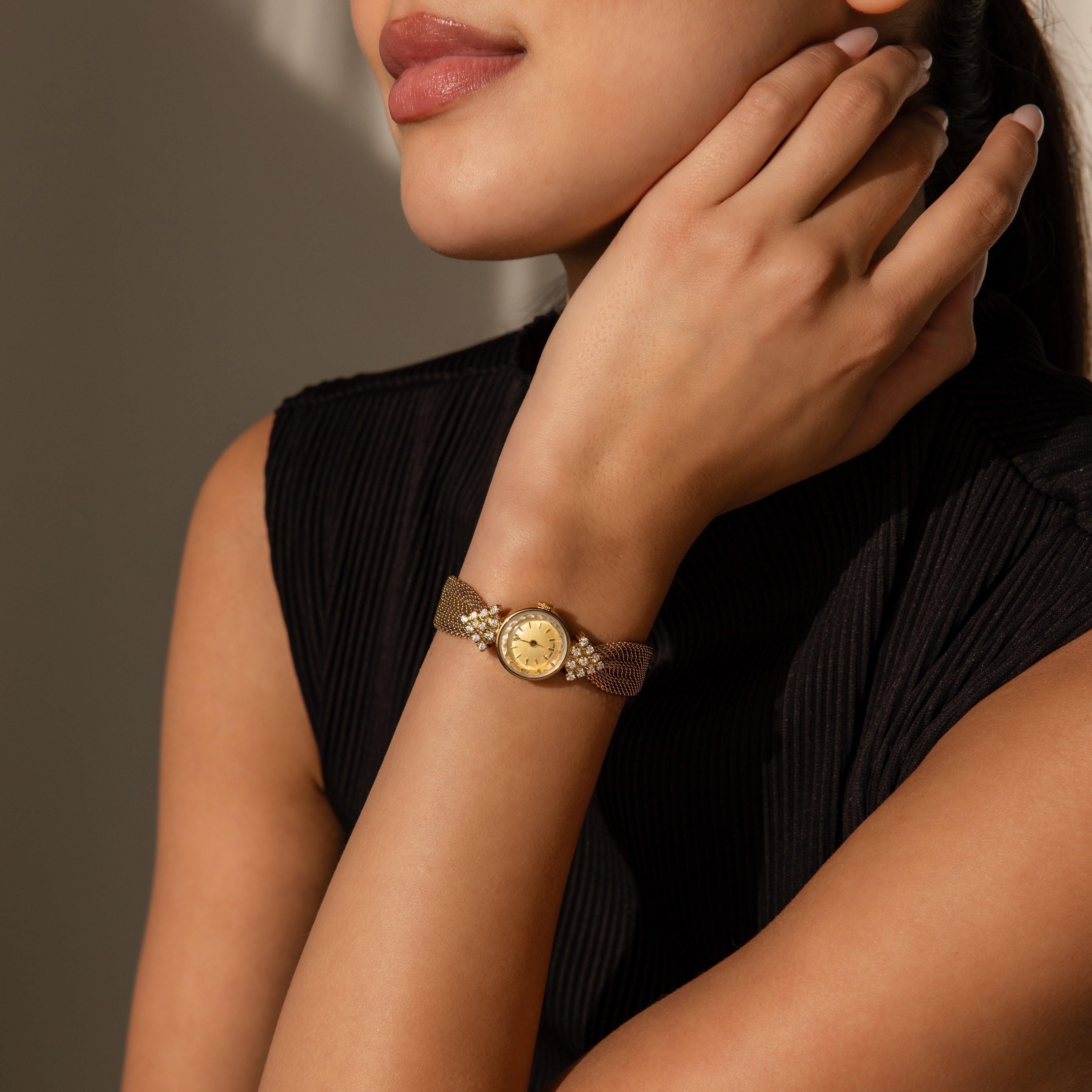 A woman wearing the Diamond Mesh Timepiece with a mesh band gently touches her neck, dressed in a black sleeveless top.