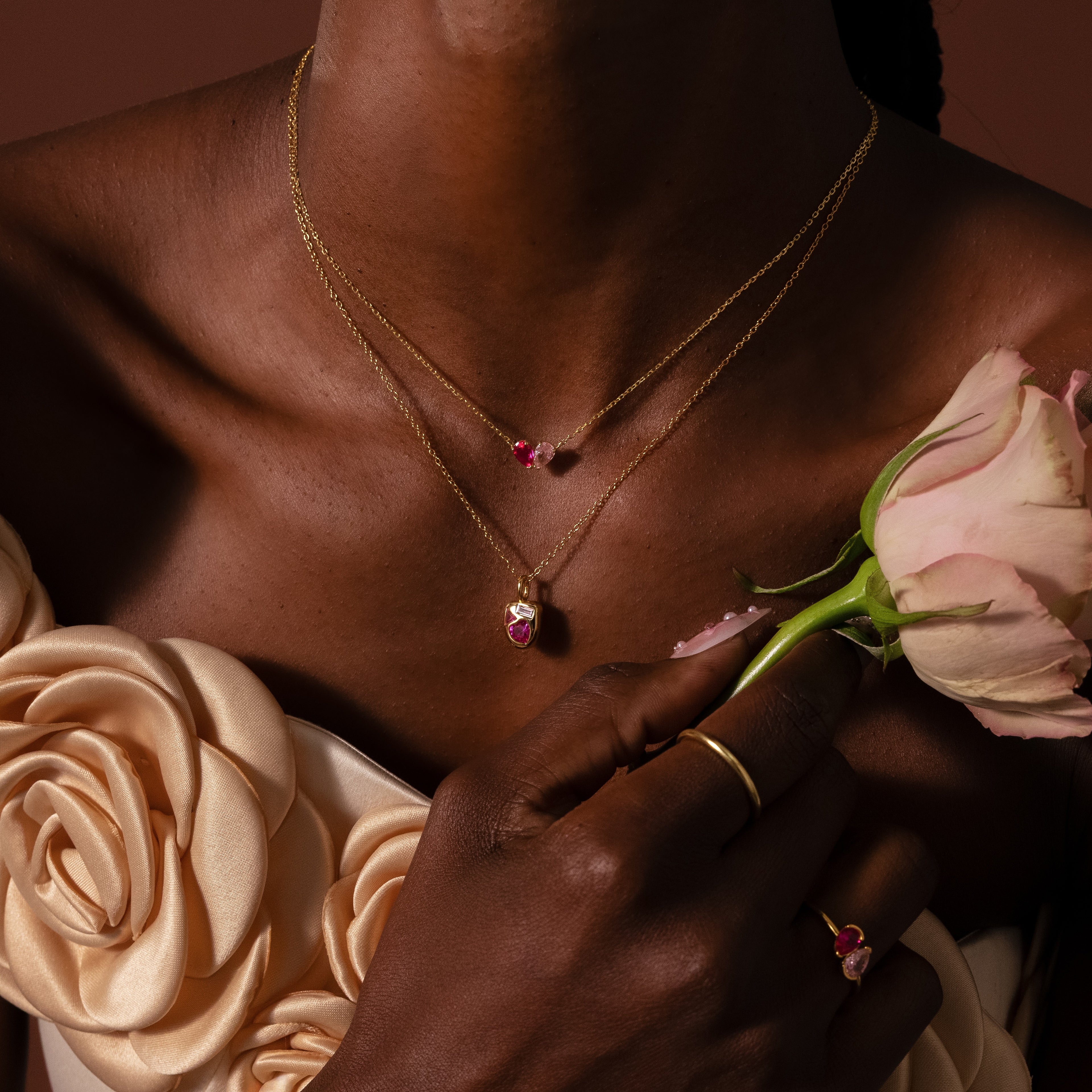 Woman in a cream floral dress holding a pink rose, wearing gold jewelry with pink gemstones and the Toi et Moi Teardrop Heart Necklace in 18K Gold.