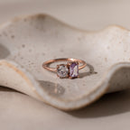 The Toi et Moi Emerald Birthstone Ring with a clear round gem and a purple rectangular gem is displayed on a textured ceramic dish.