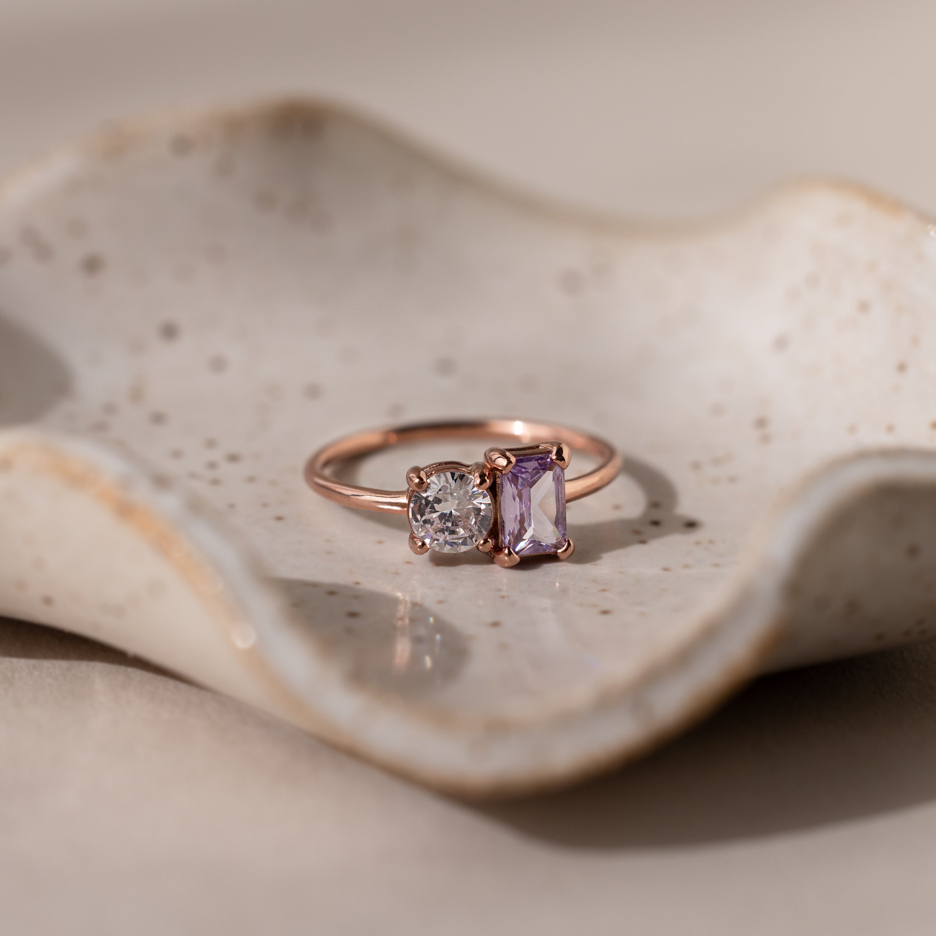 The Toi et Moi Emerald Birthstone Ring with a clear round gem and a purple rectangular gem is displayed on a textured ceramic dish.