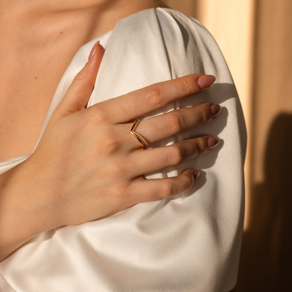 A hand with manicured nails wears the Curve Stacking Set of thin gold bands, resting on a white satin sleeve in soft light.