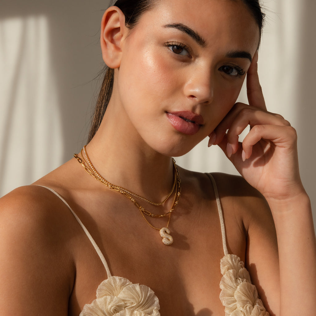 Woman wearing layered gold necklaces, including the Pave Puff Letter Necklace in 18K Gold, styled with a cream floral top and posing with her hand near her face.