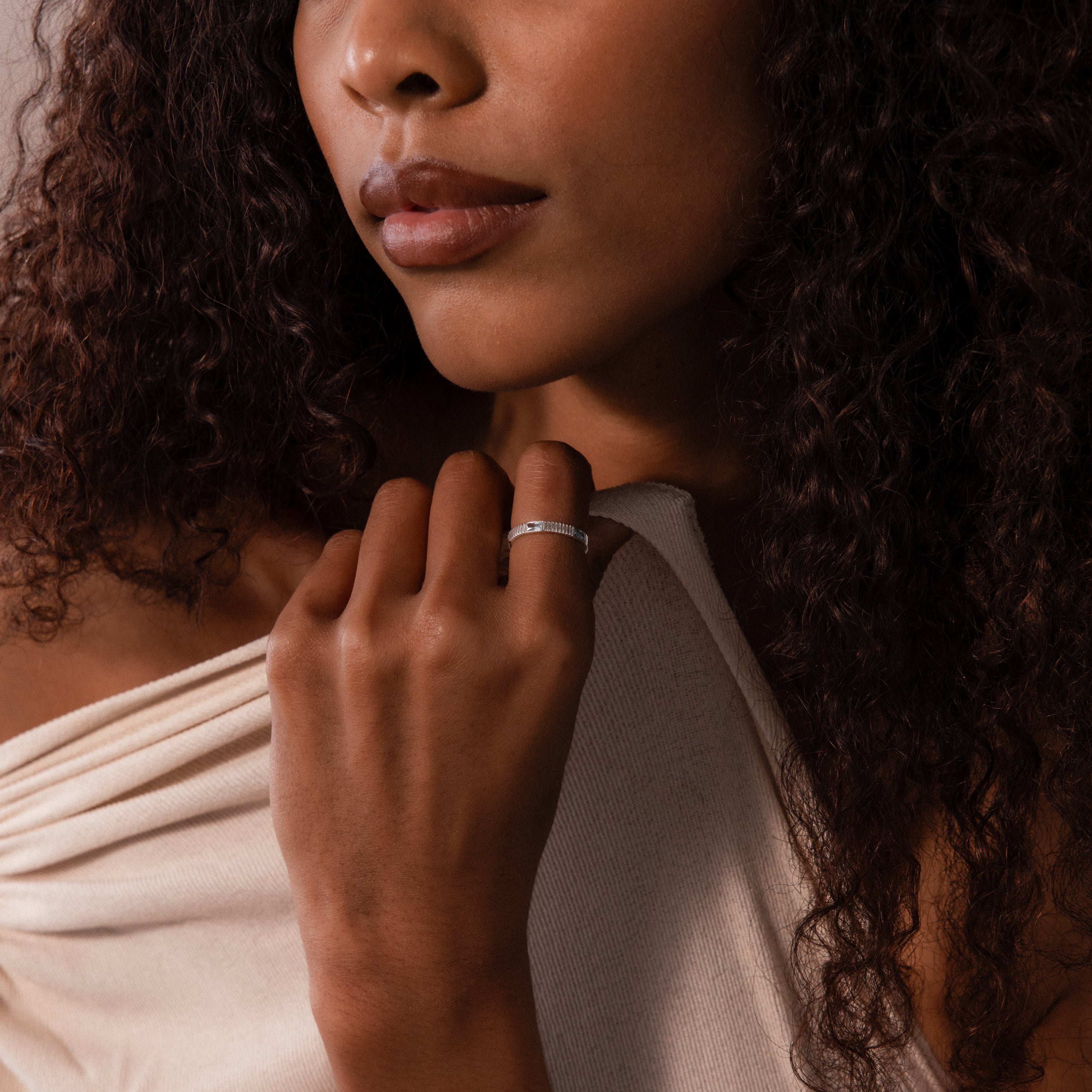 Woman with curly hair wearing an off-shoulder top, hand to her chin, showcases a sparkling Ribbed Baguette Birthstone Eternity Ring.