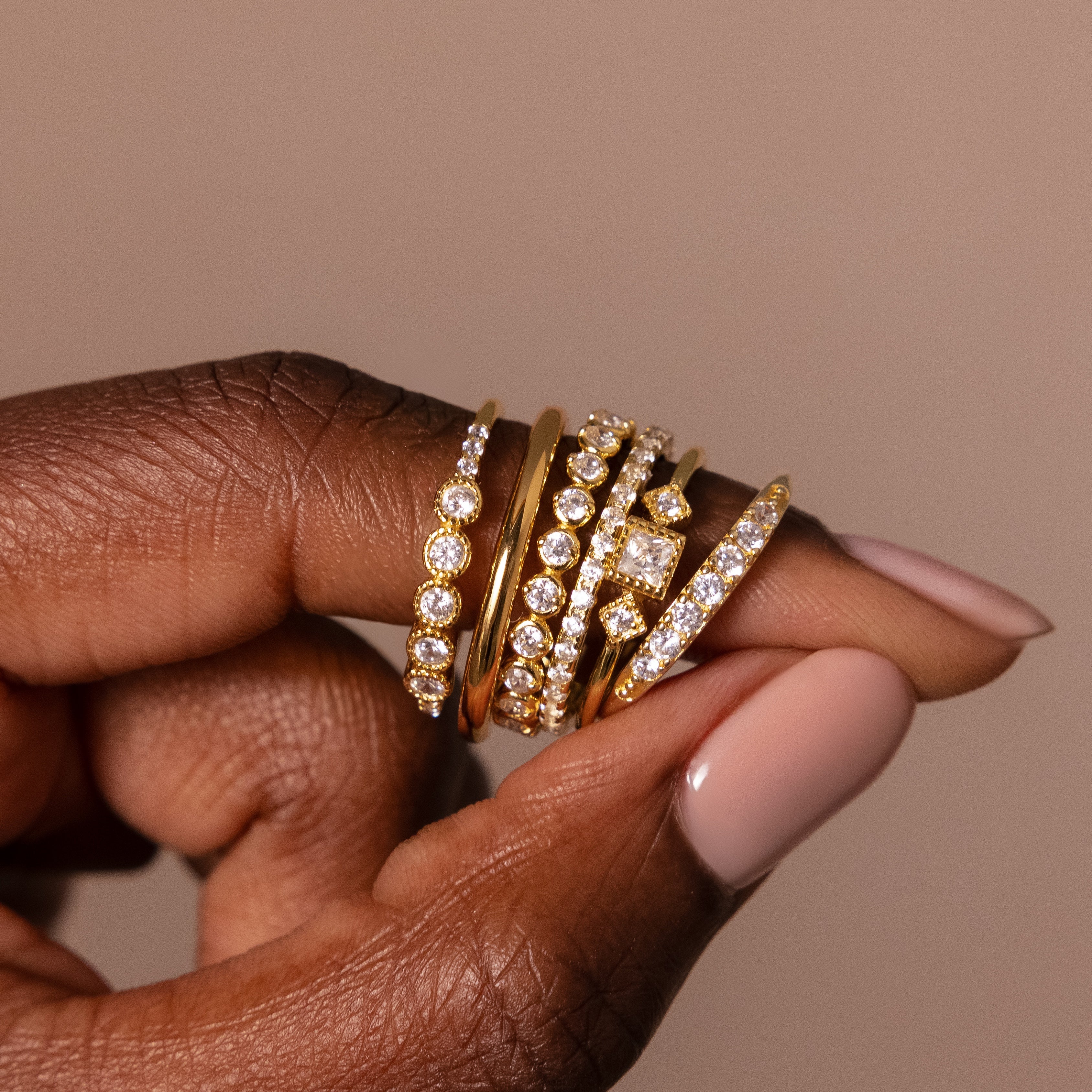A hand with nude polish displays five Art Deco Diamond Rings, each set in 18K gold and adorned with sparkling clear gemstones, exuding vintage glamour and elegance.