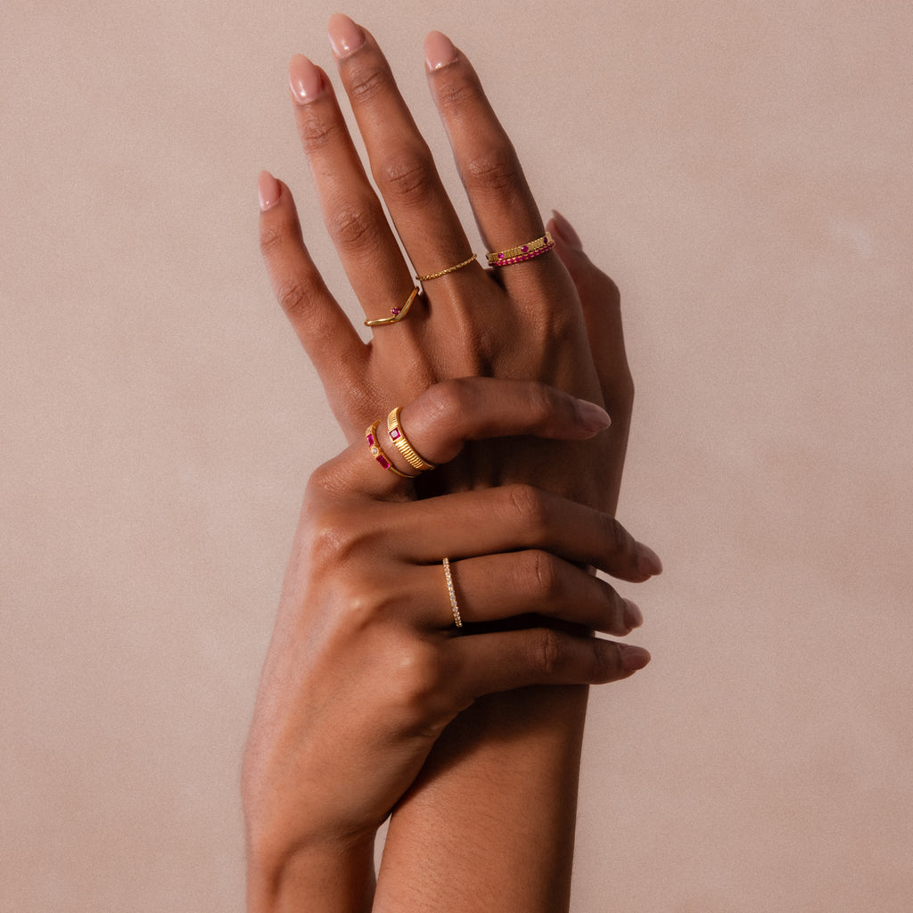 Two hands with manicured nails display multiple gold rings, including the Ribbed Garnet Band, against a neutral background.