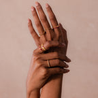 Two hands with manicured nails display multiple gold rings, including the Ribbed Garnet Band, against a neutral background.