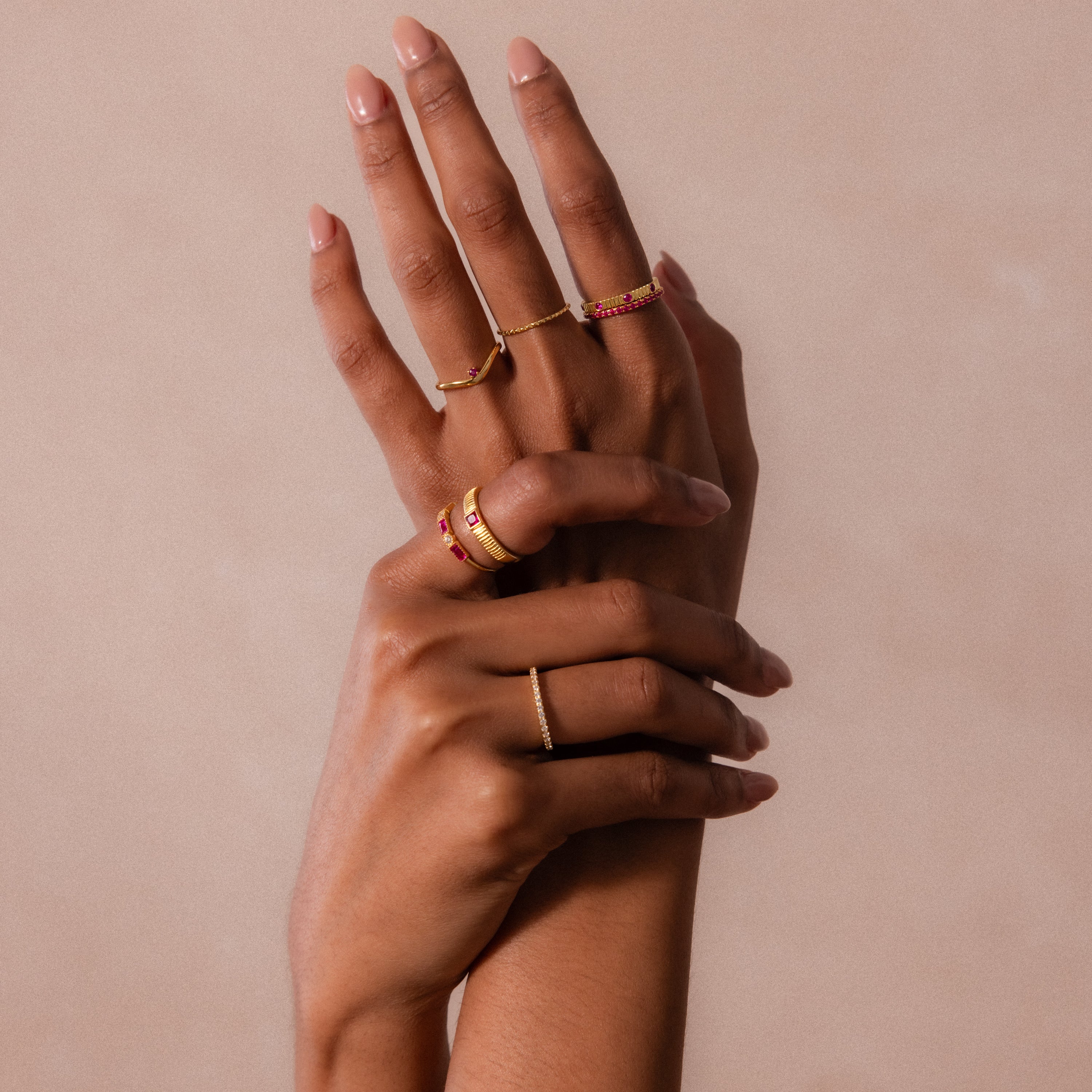 Two hands with manicured nails display multiple gold rings, including the Ribbed Garnet Band, against a neutral background.